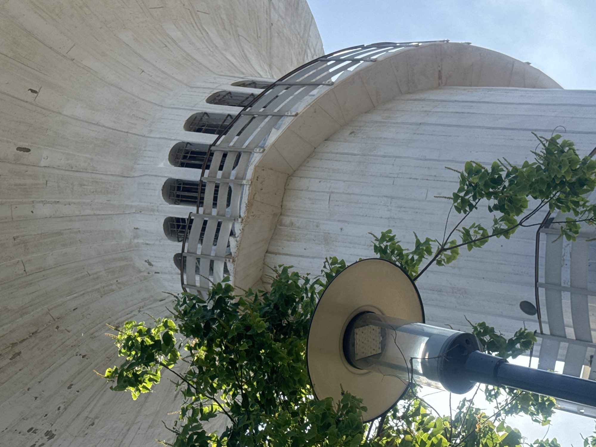 The image shows a low-angle view of the water tower in Ramat Gan, Israel, on a bright, clear day. The white, cylindrical structure dominates the frame, with its curved, fluted surface extending upwards. A circular balcony wraps around the tower, featuring a metal railing and a series of arched windows with dark interiors visible behind bars. Lush green foliage from a nearby tree partially obscures the lower section of the tower and a street lamp in the foreground. The street lamp has a wide, conical shade and a clear cylindrical housing containing a modern LED light fixture. The sky is a pale blue, with hints of sunlight filtering through the leaves and reflecting off the lamp.