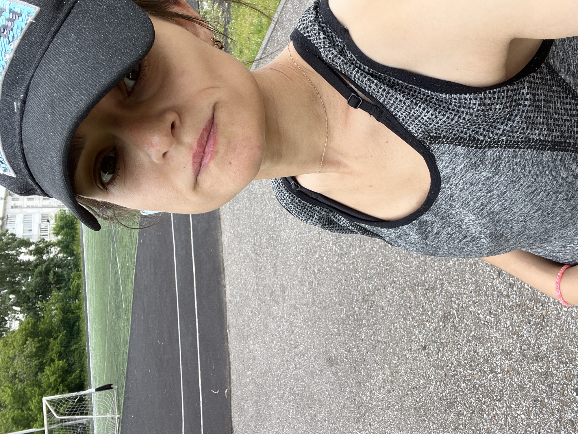 A medium shot, rotated 90 degrees clockwise, captures a person, likely female, taking a selfie outdoors at an athletic facility in Chaikovsky, Russia. The individual looks directly at the camera with a neutral to slightly content expression.
She wears a dark-colored cap or visor, which features light blue or silver sequin-like details on its side. Her athletic attire consists of a grey and black patterned sports top or tank top with black trim. A thin, delicate gold-colored chain necklace is visible around her neck, and a pink beaded or woven bracelet adorns her right wrist.
The background clearly depicts an outdoor sports environment. To the left, a green artificial turf soccer field with a white soccer goal net is visible. To the right, a dark grey asphalt running track with prominent white lane lines stretches out. Beyond the field and track, lush green trees and distant residential or commercial buildings can be seen under what appears to be an overcast or diffused daylight sky. The ground in the foreground is paved with light grey aggregate, typical of walkways around sports complexes. There is no visible text. The overall impression is of a casual moment captured at an active outdoor recreational area.