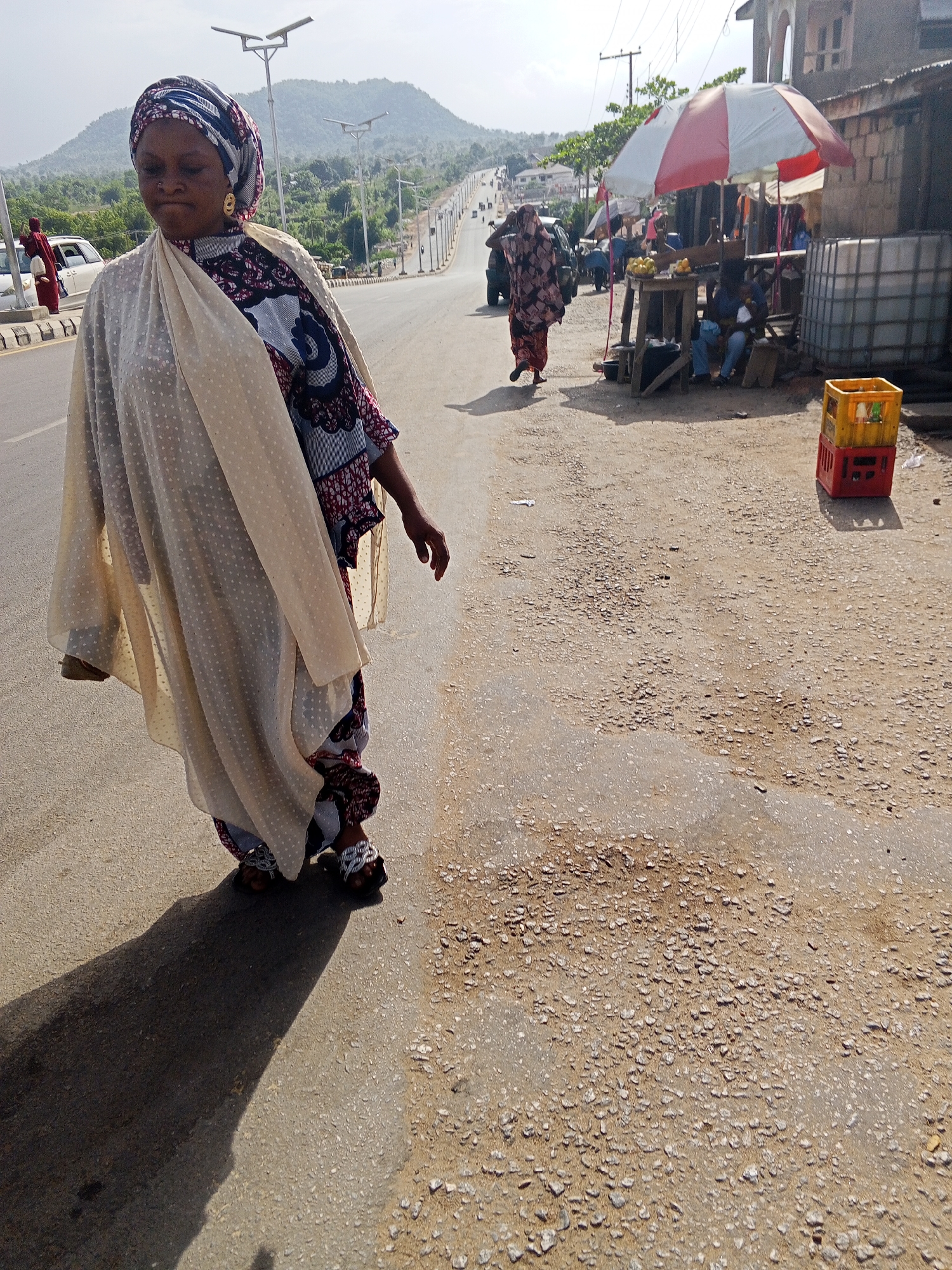 The image depicts a sunlit roadside scene in Adankolo, Nigeria. In the left foreground, a woman walks, casting a long shadow. She is dressed in a colorful headwrap, a patterned garment, and a sheer, light-colored overlay with small white dots. She wears golden hoop earrings and embellished sandals. Her eyes are lowered, and her expression appears neutral or contemplative.
Further down the road in the mid-ground, another person in patterned attire walks away from the camera. On the right side of the frame, a street vendor's stall is set up on the dirt shoulder, shaded by a prominent red and white striped umbrella. What appears to be fruit, possibly oranges, is displayed on a wooden stand. Several individuals are seated under the umbrella, suggesting activity at the stall. Large white water storage containers are visible next to the vendor setup, and a stack of plastic crates—a yellow one atop a red one—rests on the ground.
The road, which appears to be paved, stretches into the distance and is lined with modern streetlights, each equipped with a solar panel. In the background, green hills rise under a bright, clear sky. The ground transitions from paved road to a rough, gravelly dirt shoulder. The long shadows cast by the woman and other objects indicate either early morning or late afternoon. No discernible text is clearly legible on any of the objects.