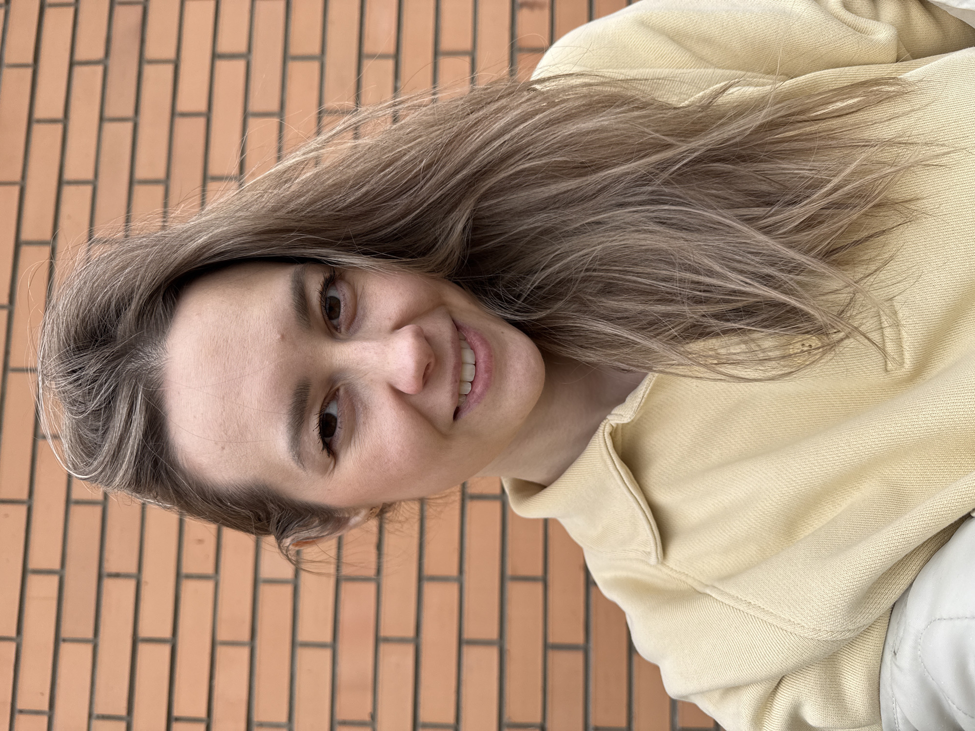 A woman with long blonde hair and brown eyes smiles at the camera. She is wearing a yellow sweatshirt. The background is a brick wall. There is no specific information available to determine the location is in Chaikovsky, Russia.