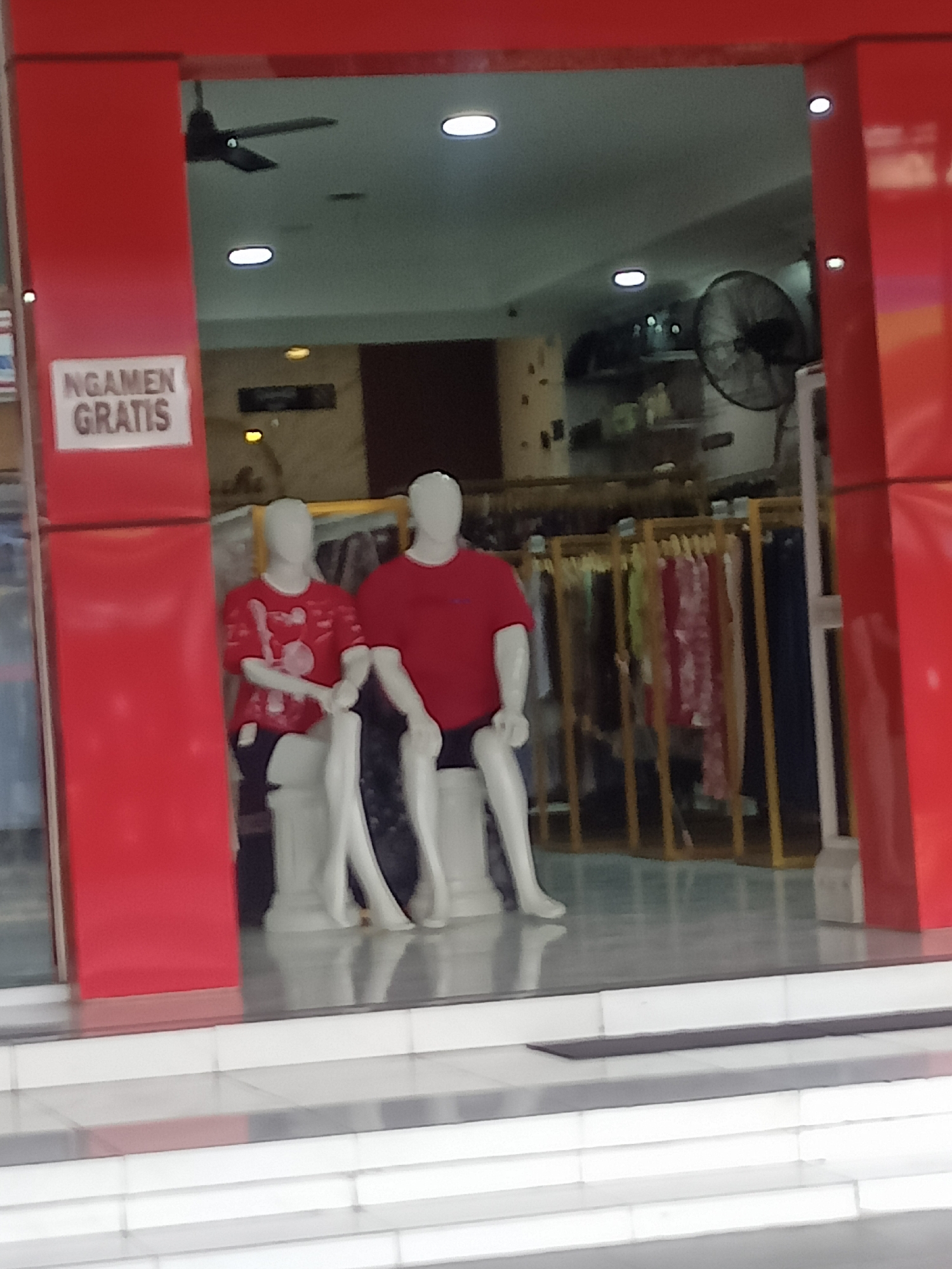 The image shows a storefront in Gemolong, Indonesia. A red pillar is on the left side of the image, with a sign that reads "NGAMEN GRATIS." In front of the storefront, there are two mannequins dressed in red t-shirts and dark shorts. Behind the mannequins, there are racks of clothing for sale. A ceiling fan is visible on the ceiling. The store appears to be a clothing store.