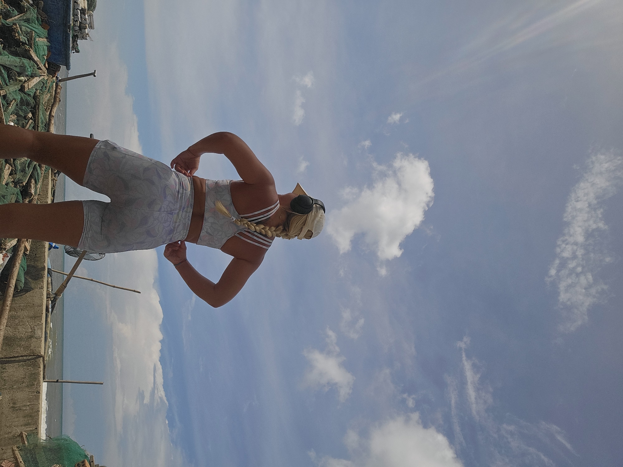A blonde woman wearing a white baseball cap, headphones, and a matching white patterned sports bra and shorts stands with her back to the camera. She is on a concrete embankment overlooking the ocean in Mũi Né, Vietnam. Piles of fishing nets and debris are visible on the embankment. The sky is blue with scattered white clouds.