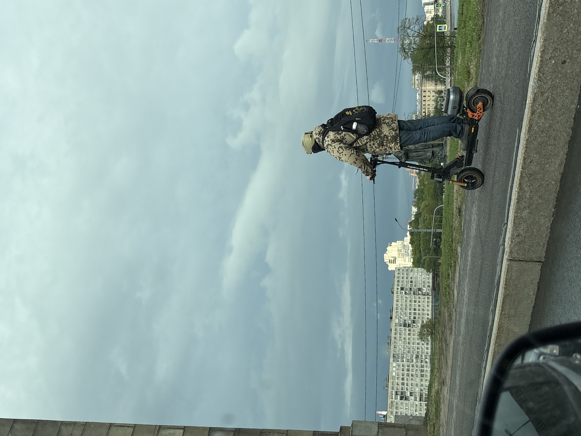 A medium shot reveals a person riding an electric scooter on an urban road, the scene framed by a cloudy sky above and building structures in the background. The rider, facing left, is outfitted in a camouflage jacket, blue jeans, and a helmet, and wears a black backpack. The scooter itself is black with orange accents. The backdrop includes a high-rise building with numerous windows and a crane visible in the distance. The image was likely taken from inside a car. The location is Saint Petersburg, Russia.