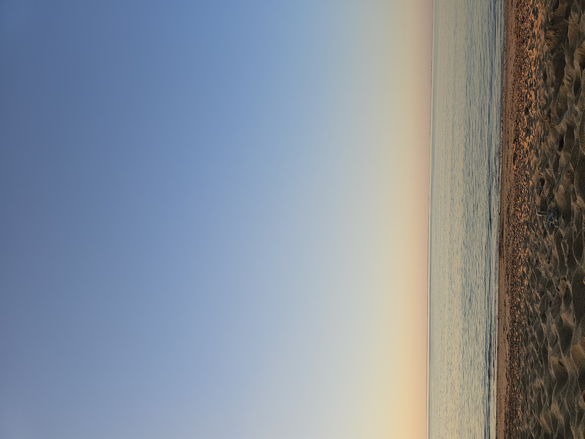 The image depicts a beach scene in La Brée-les-Bains, France. The left portion of the frame showcases a gradient sky transitioning from a light blue at the top to a pale yellow-orange towards the horizon. The right portion features the sea, a narrow strip of shoreline with pebbles, and a sandy beach. The sand shows textures and patterns indicative of footprints or raking. The water is calm, with subtle wavelets visible near the shoreline. No people or objects are visible in the sea, on the beach, or in the sky.