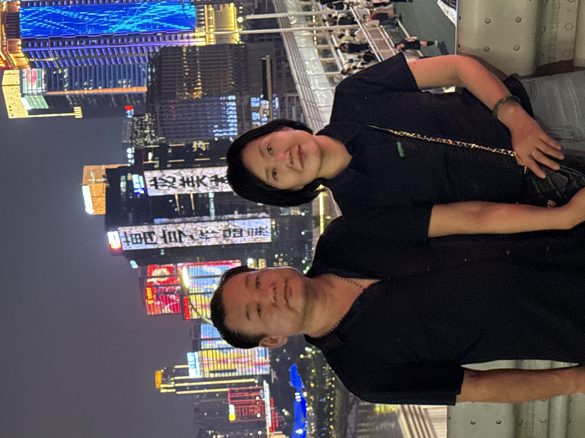 A man and a woman stand in front of a nighttime cityscape in Yuzhong District, China. The man, on the left, wears a black collared shirt and a necklace. The woman, on the right, wears a black collared shirt and a purse with a chain strap. Both individuals are looking towards the camera. Behind them, illuminated skyscrapers dominate the background, with neon lights and advertisements displayed on their facades. A bridge or elevated walkway with railings is visible in the mid-ground, with numerous people walking on it. The overall scene depicts a vibrant urban environment at night.