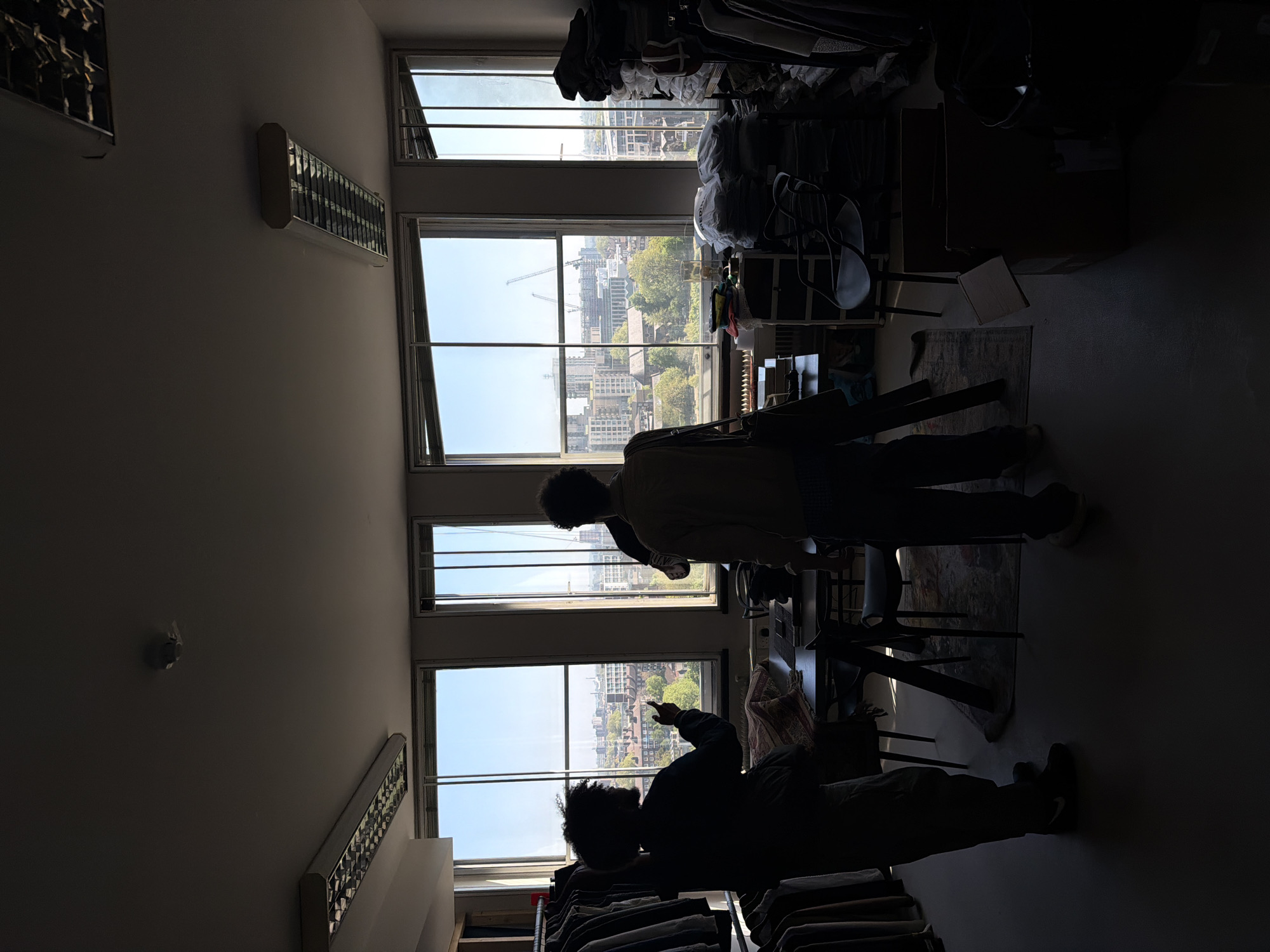 The image depicts the interior of a room with two individuals silhouetted against a set of windows revealing a cityscape, likely London, United Kingdom. The windows show buildings, including what appears to be cranes from construction sites, and greenery suggesting parks or trees within the city. One person is pointing towards the outside, suggesting an engagement with the view. Clothes racks and other furniture clutter the room. Two HVAC units are mounted on the wall. A smoke detector is visible on the ceiling. The room appears dimly lit, with the outside daylight serving as the primary light source.