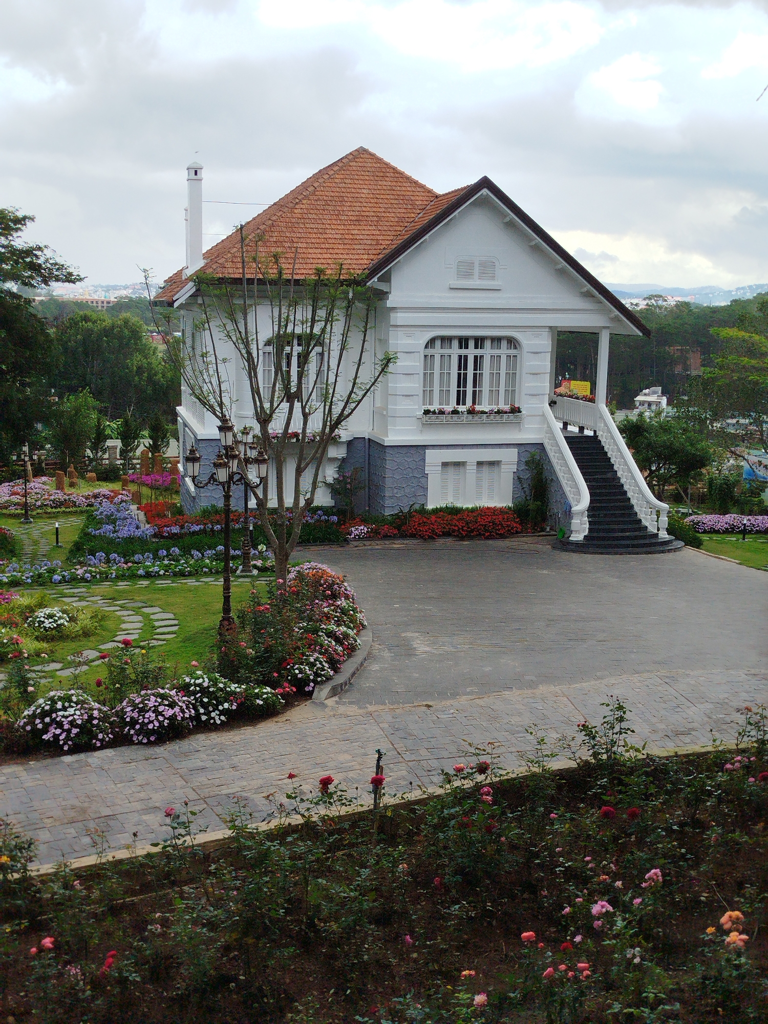 The image shows a white two-story house with a brown tiled roof in Da Lat, Vietnam. The house has a small porch with stairs leading up to the second floor. The lower level is painted a light blue, while the upper level is white. The surrounding landscape includes manicured gardens with various flowers, a stone-paved driveway, and a green lawn with a stone pathway. In the background, there are trees and a cityscape under a cloudy sky. There are no visible people in the image.