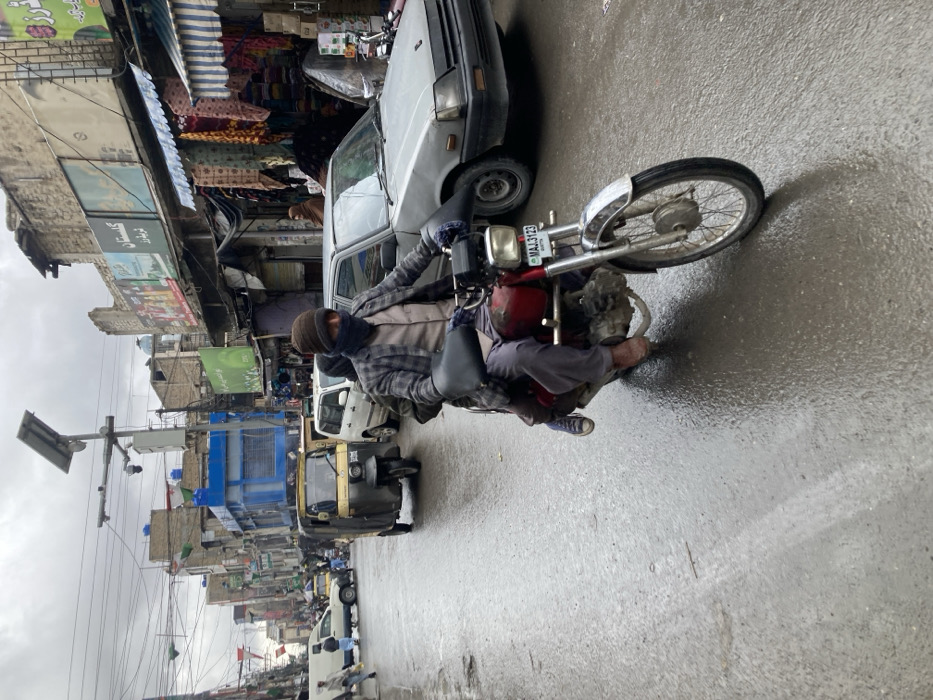 This image depicts a wet urban street scene in Quetta City Tehsil, Pakistan, under an overcast sky. A red motorcycle with registration plate MAJ 3123 is centrally featured, moving from the right foreground towards the left. The driver is a male wearing a dark brown beanie, a dark neck gaiter, a plaid jacket, and gloves. A second person, partially obscured, is seated behind the driver, wearing grey trousers and blue sneakers.
To the left of the motorcycle, a yellow auto-rickshaw is visible, followed by a white van and other distant vehicles. On the right, a silver sedan is parked alongside the road. The street surface is visibly wet, reflecting light, indicating recent rain. Buildings line both sides of the street, featuring multi-story structures with varying architectural styles, some appearing aged or partially damaged. Commercial storefronts are present, displaying awnings, merchandise, and signage primarily in Urdu/Persian script. Overhead, numerous utility lines crisscross the street, and a pole equipped with a solar panel and multiple CCTV cameras is prominent. Several individuals are visible walking on the street's edges in the background and midground.