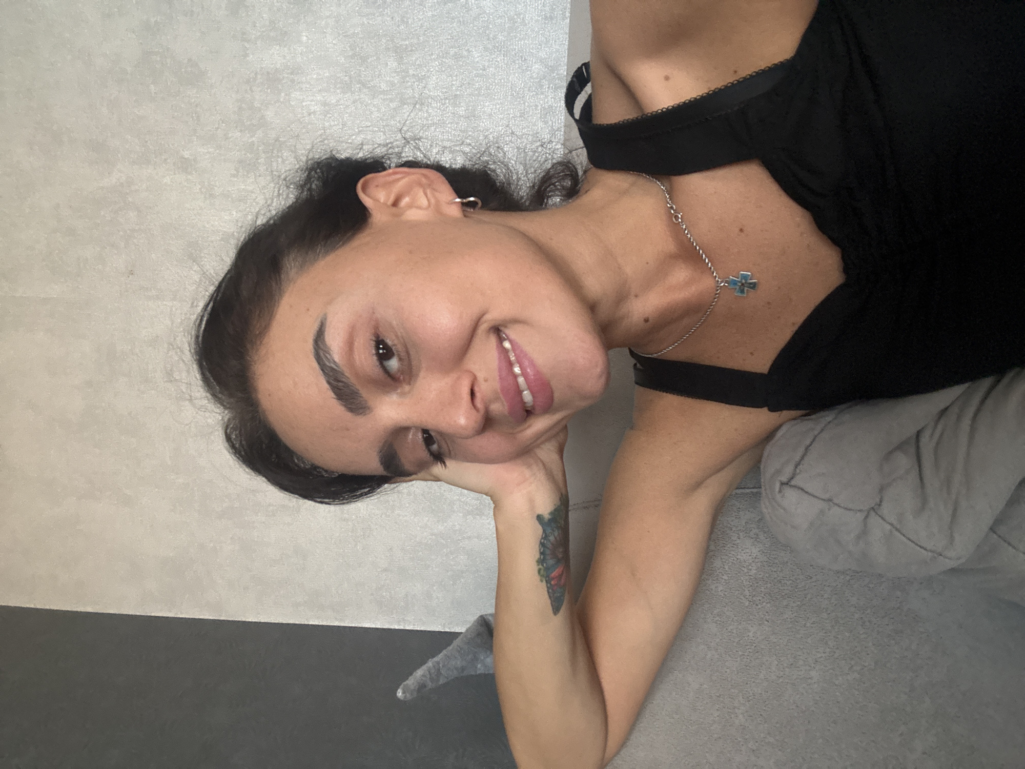 In Tbilisi, Georgia, a young woman with dark hair is relaxing on a sofa, leaning her head on her hand. She is wearing a black top with thin straps and a silver chain with a cross pendant. She has a tattoo on her arm and is smiling. The background appears to be a wall with a textured pattern.