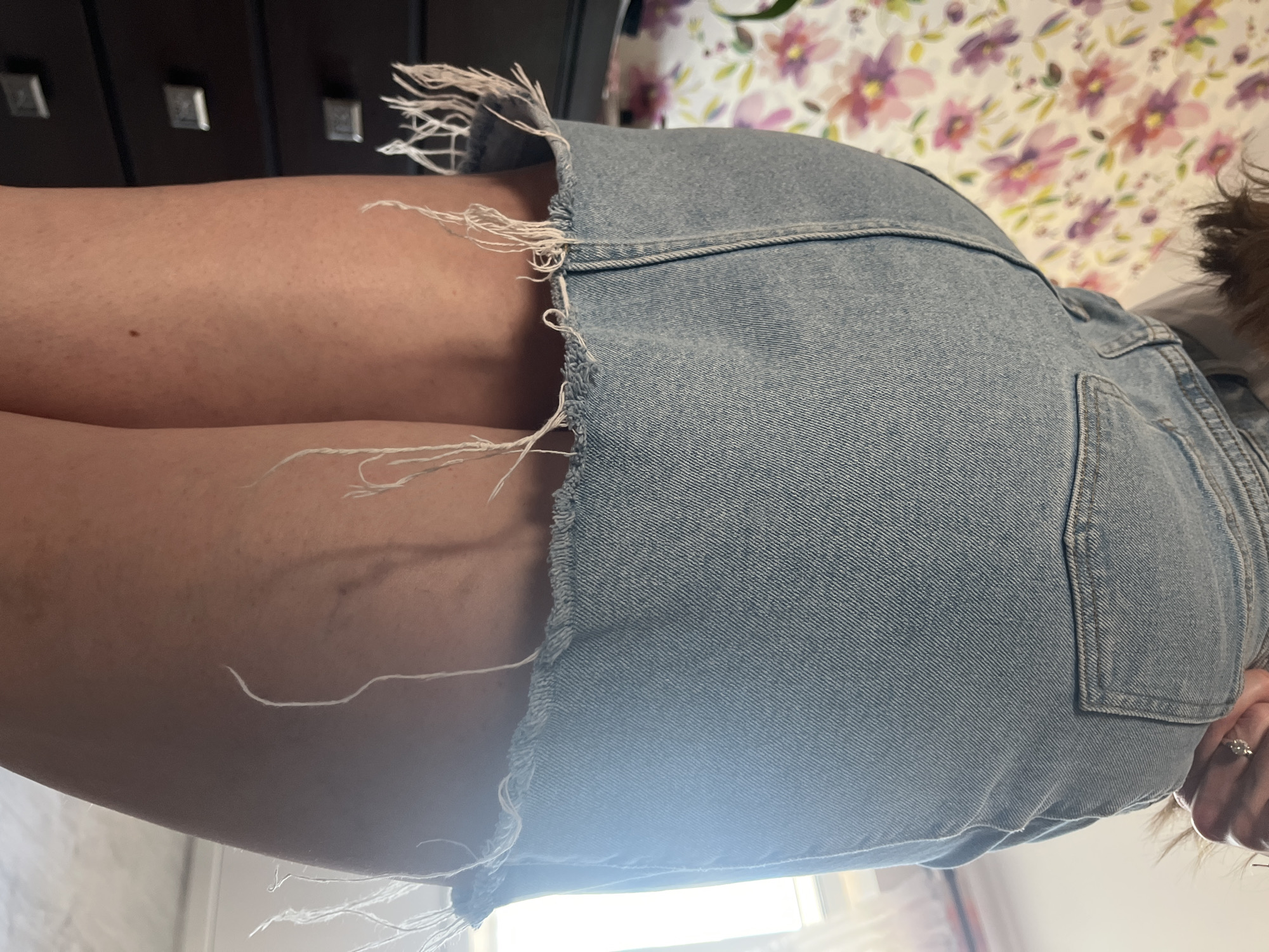 The image shows a close-up view of a person's legs and a denim skirt. The skirt is light blue denim with a frayed hem and loose threads hanging down. The person's skin is visible around the skirt, and appears to have some faint vein lines. In the background, a dark piece of furniture with square handles is visible, possibly a dresser or cabinet. Further in the background, a floral patterned wallpaper with pink and purple flowers can be seen. The lighting suggests it could be daytime, possibly indoors, given the presence of furniture and wallpaper. There is no text visible in the image. The image is an angled shot, focusing on the texture of the denim and the skin.