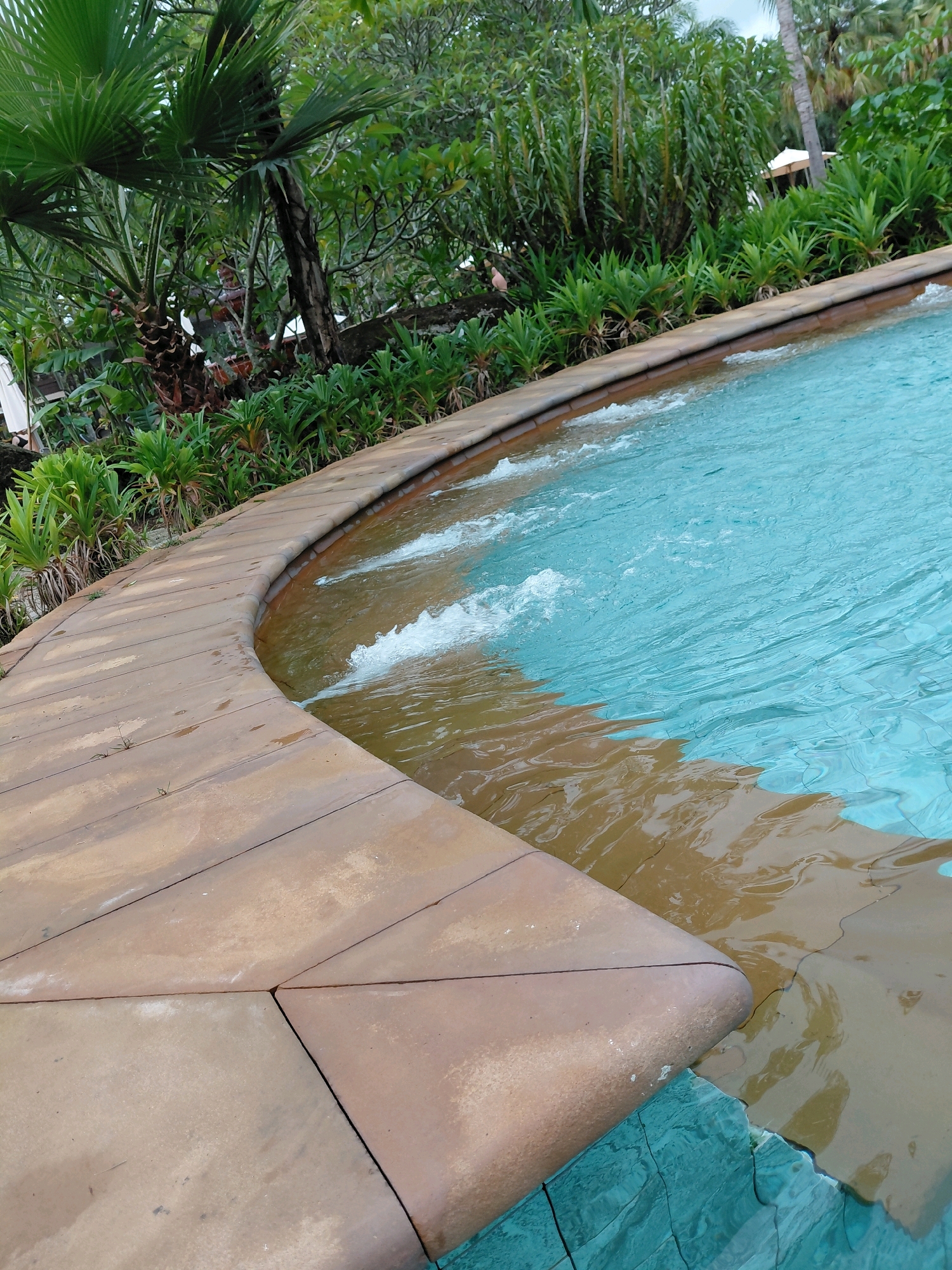 The image depicts a portion of a swimming pool with turquoise water and a curved, tan-colored tiled edge. The water is rippling, indicating movement, and there are several small waves or splashes visible near the edge. Lush green foliage, including palm trees and other tropical plants, forms a dense backdrop to the pool area. In the distance, a white umbrella or canopy is partially visible, suggesting it is an outdoor setting, likely a resort or hotel. The scene is located in Ban Khuan, Thailand.