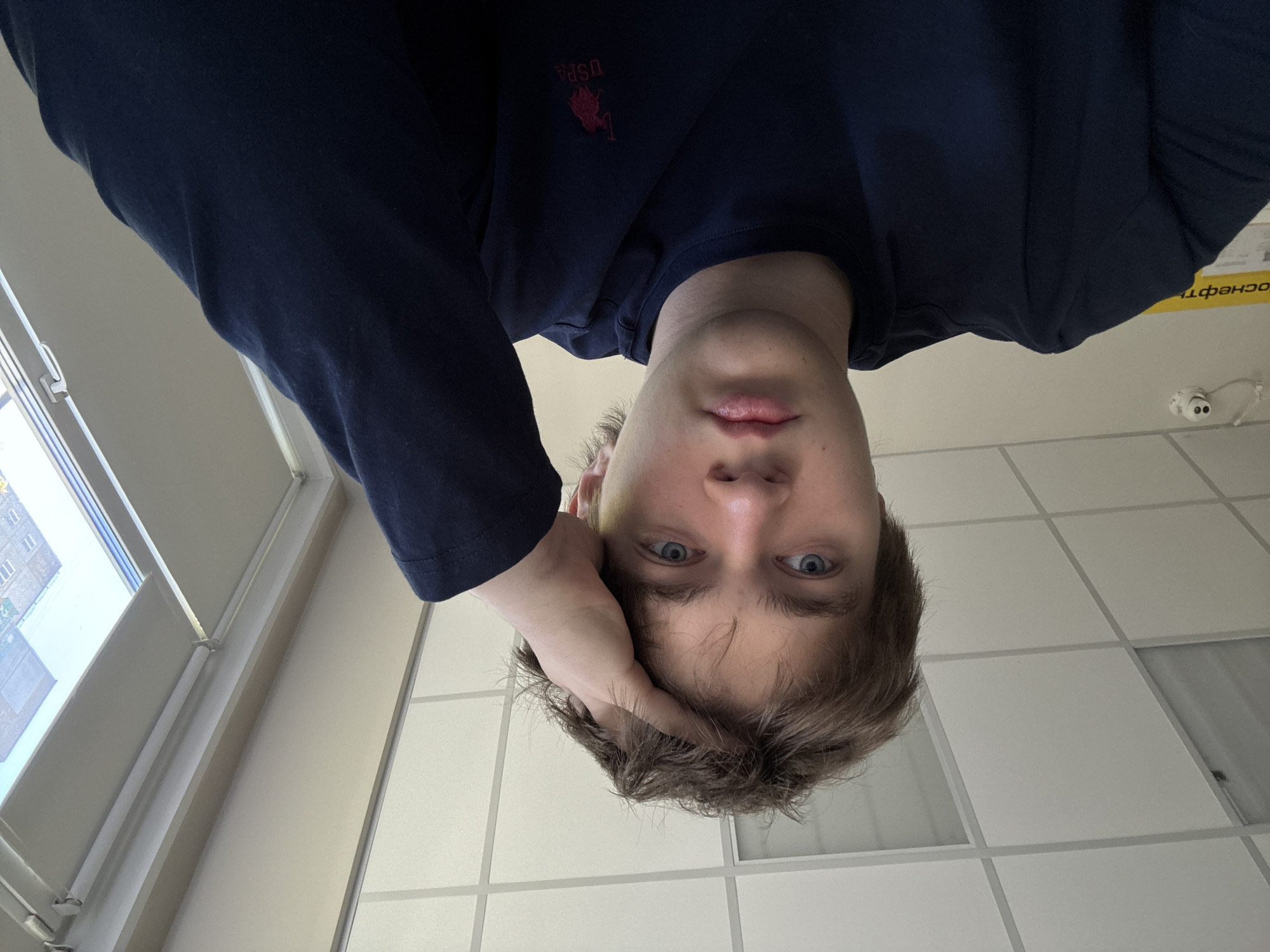 A young man is looking down into the camera, his right hand touching his head. He is wearing a dark blue long-sleeved shirt with a "USPA" logo. The background consists of a white tiled ceiling, a window to the left showing an exterior view of buildings, and a yellow sign on the wall in the top right corner with Russian writing (likely related to "Rosneft" based on the visible Cyrillic letters). The location is likely in Krasnoyarsk, Russia, inferred from the Russian text and likely presence of Rosneft gas stations in the area.