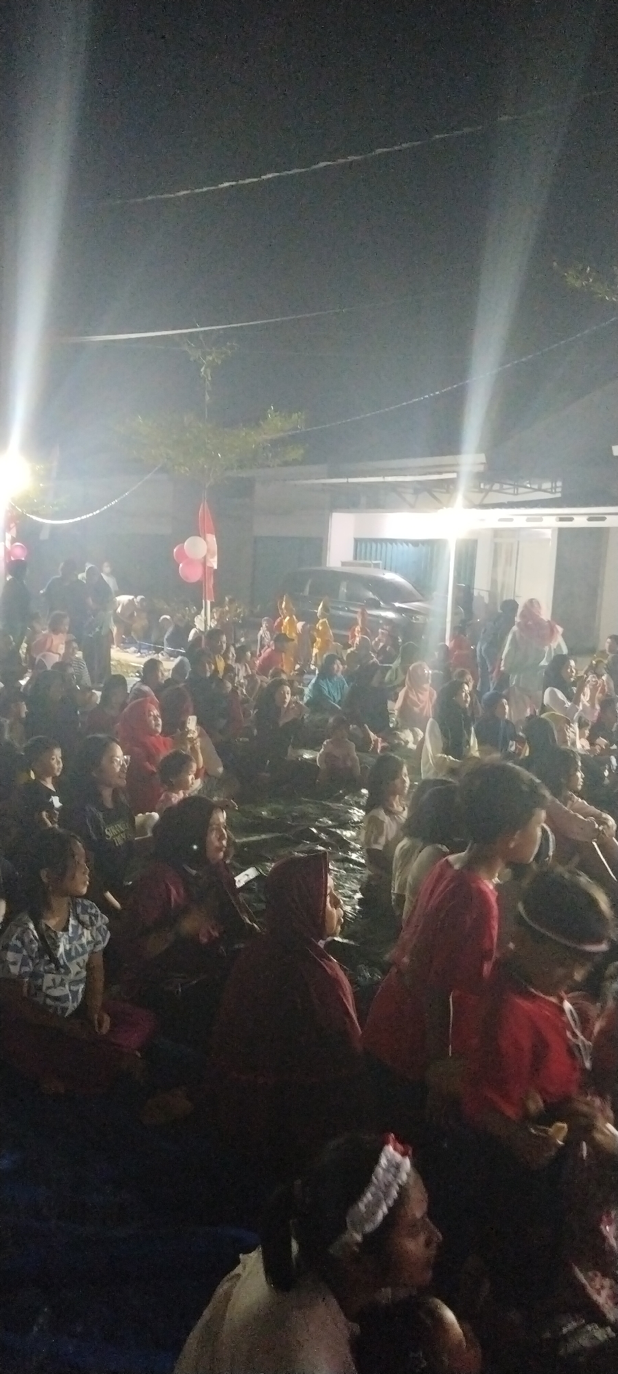 This is a low-angle, full shot taken at night of a crowd of people, likely attending an outdoor event. The event appears to be taking place in a residential or semi-public outdoor space, possibly in Taban, Indonesia, given the context. The lighting is dim, with bright spotlights casting long beams of light downwards, creating a somewhat hazy atmosphere. The ground is covered with dark blue tarps.
In the foreground and middle ground, the crowd is seated or kneeling on the tarps, facing towards the right of the frame, suggesting they are watching a performance or event in that direction. Many individuals are wearing headscarves, indicating a likely Muslim population. Various clothing colors are visible, including reds, burgundies, blues, and whites. Some children are present within the crowd.
In the background, there are buildings with white walls and darker roof structures. A black car is parked in front of one of the buildings. A tree with sparse foliage is visible between the crowd and the buildings. Further back, there are more figures and structures that are less distinct due to the distance and lighting conditions. A string of lights or decorations is visible above the crowd, connecting to poles on either side. Three balloons, one red and two white, are clustered on a pole near the center-left of the image. The overall impression is of a community gathering or celebration.