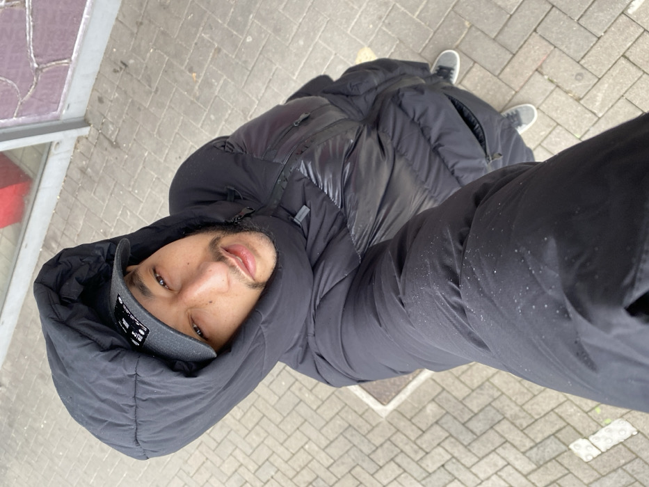 An adult male is shown in a high-angle, downward-facing self-portrait taken outdoors in Bischheim, France. The subject, with a beard, wears a black hooded puffer jacket with the hood up, and a dark grey baseball cap beneath it, featuring a white logo. Small white specks are visible on the left jacket sleeve. His lower body, clad in dark trousers and grey sneakers, is partially visible. The ground is paved with light grey rectangular interlocking pavers. In the background, sections of a building are visible, featuring a glass panel, a red surface, and a textured purple-brown wall.