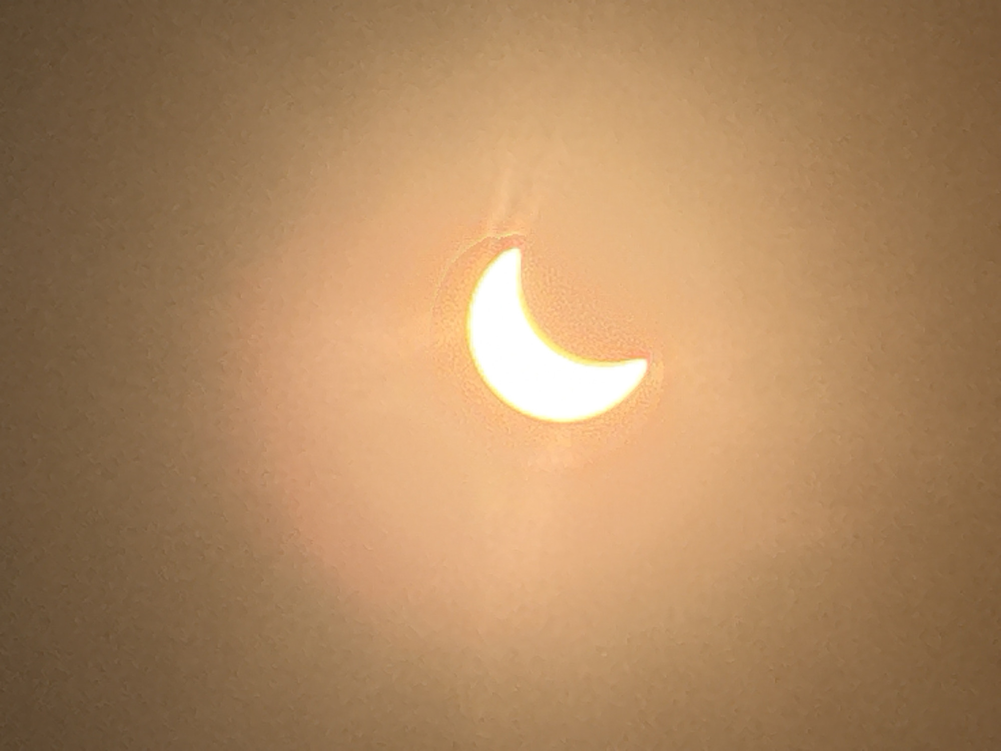 A crescent shape of bright white light, representing a solar eclipse, is centered in the frame against a muted, textured background of warm, earthy tones, likely representing a ceiling or wall illuminated by ambient light. The crescent is positioned slightly to the left of the vertical center. The light source appears to be a projection or a captured image of a solar eclipse, given the distinct crescent shape and the halo effect around it. The background exhibits a subtle, speckled texture, indicating a non-uniform surface. There is a soft glow emanating from the crescent, diffusing into the surrounding area. The image appears to be taken indoors, with the lighting suggesting an overcast day or a controlled indoor environment where the eclipse is being viewed indirectly, possibly through a projection. There are no people, animals, or other objects visible in the image. The image was captured in Märstetten, Switzerland.