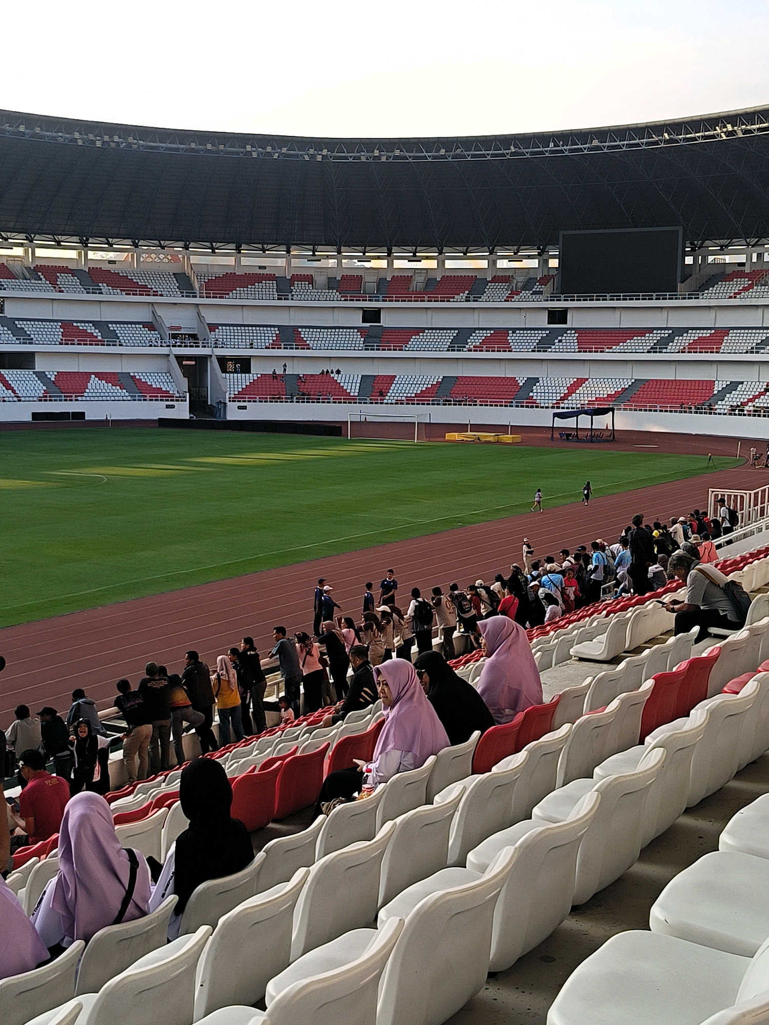 This image depicts the exterior of the Jatidiri Stadium in Semarang, Indonesia, during daylight hours. The stadium features tiered seating in red, white, and gray, arranged around a vibrant green football field with a running track. A large black screen is visible at the top of the stadium. Spectators, many wearing hijabs, are seated in the stands, observing the field. Some individuals are standing in the aisles and on the track, engaged in conversation. The overall scene suggests a sporting event or gathering at the stadium.