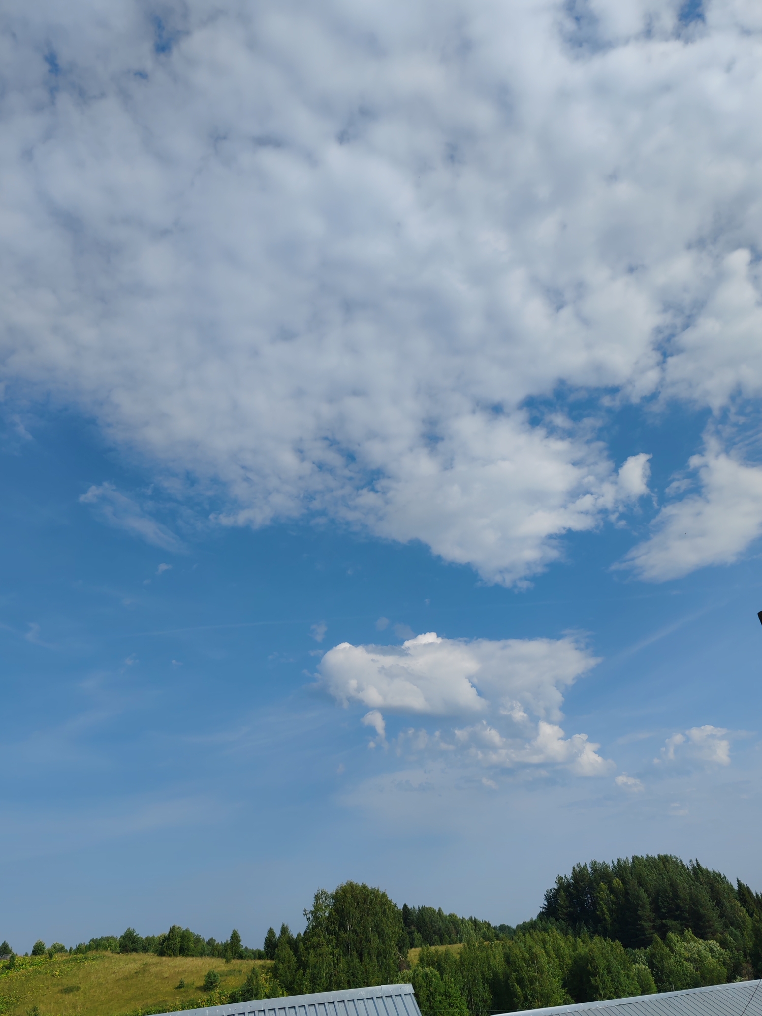 The image shows a wide shot of a rural landscape with a bright blue sky filled with fluffy white clouds. The lower half of the image consists of a rolling green hill dotted with trees and vegetation. In the foreground, there is a glimpse of a metallic roof structure. The scene appears to be a sunny day in a natural setting. The provided location of Чухлэм, Russia, suggests a rural or semi-rural environment.