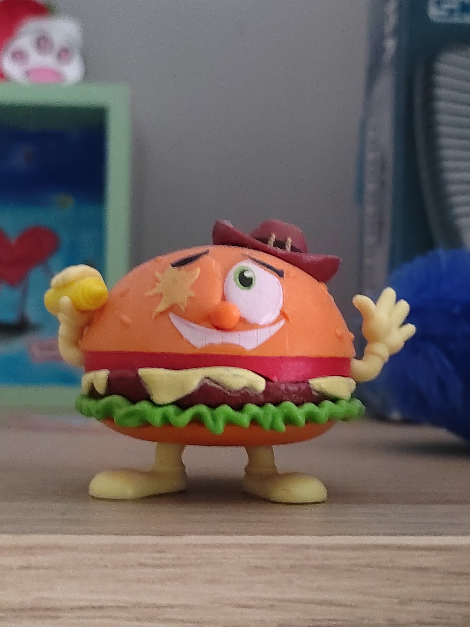 The image shows a close-up of a toy hamburger character standing on a wooden surface. The toy is a stylized hamburger with a round orange bun, red and yellow "toppings" resembling cheese and meat, and green lettuce. It has a smiling face with green eyes, an orange nose, and a wide mouth. The toy is wearing a brown cowboy hat. Its arms are outstretched, with one hand holding a small yellow object and the other waving. In the background, there is a blurry green shelf with a blue object behind it, and a fluffy blue object to the right of the toy. The environment appears to be indoors. There is no indication of the location being Porto Recanati, Italy.