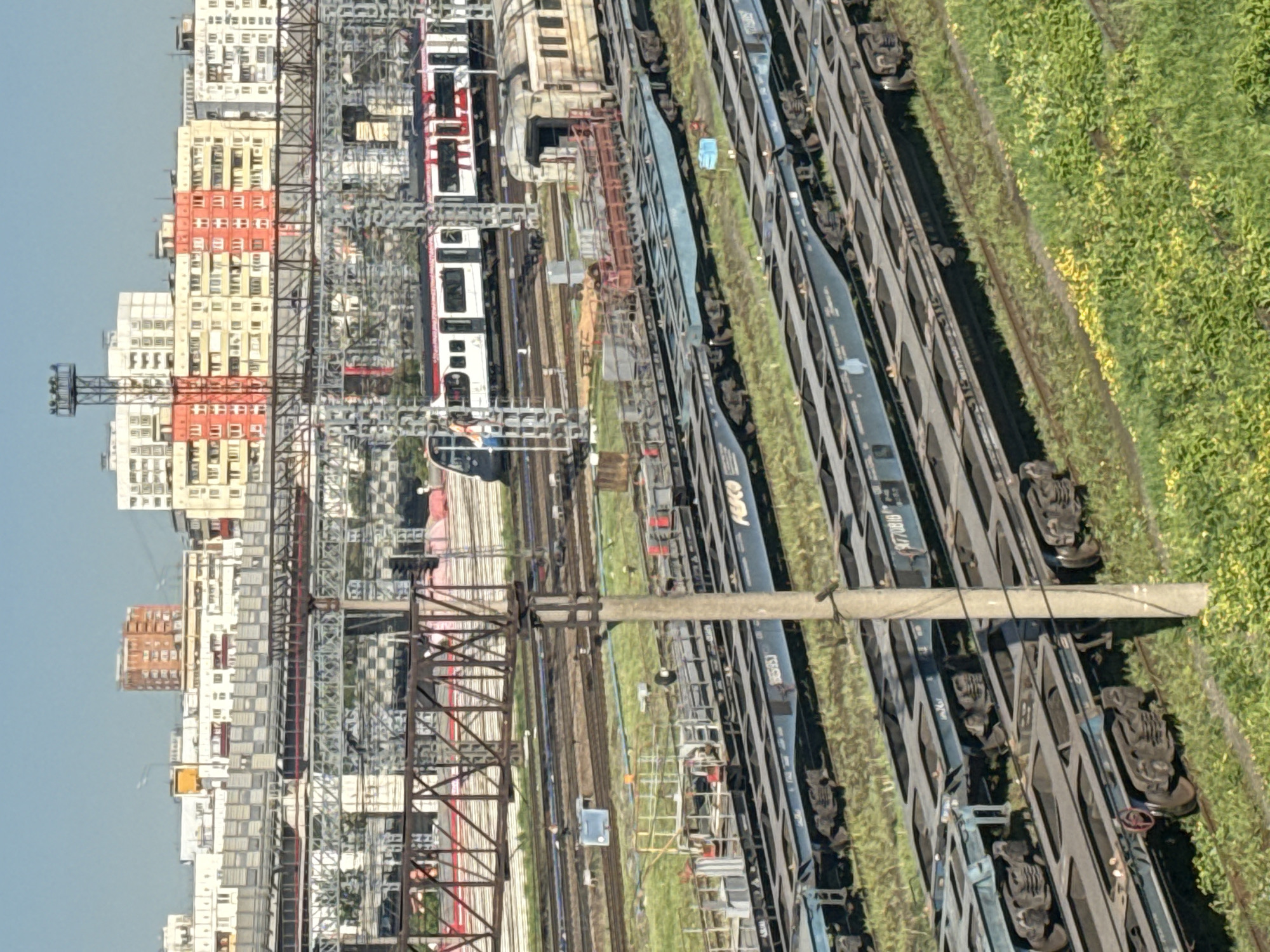 The image shows a railway yard in an urban environment, likely in Moscow, Russia, based on the architectural style of the buildings. Several parallel railway tracks run through the scene, some occupied by trains. In the foreground, there are long freight cars filled with containers. Buildings can be seen in the background, with a variety of architectural styles. Above the train tracks, a network of power lines and pylons extends. There are grassy areas and vegetation along the sides of the tracks. Some scaffolding and ladders are located near the tracks.