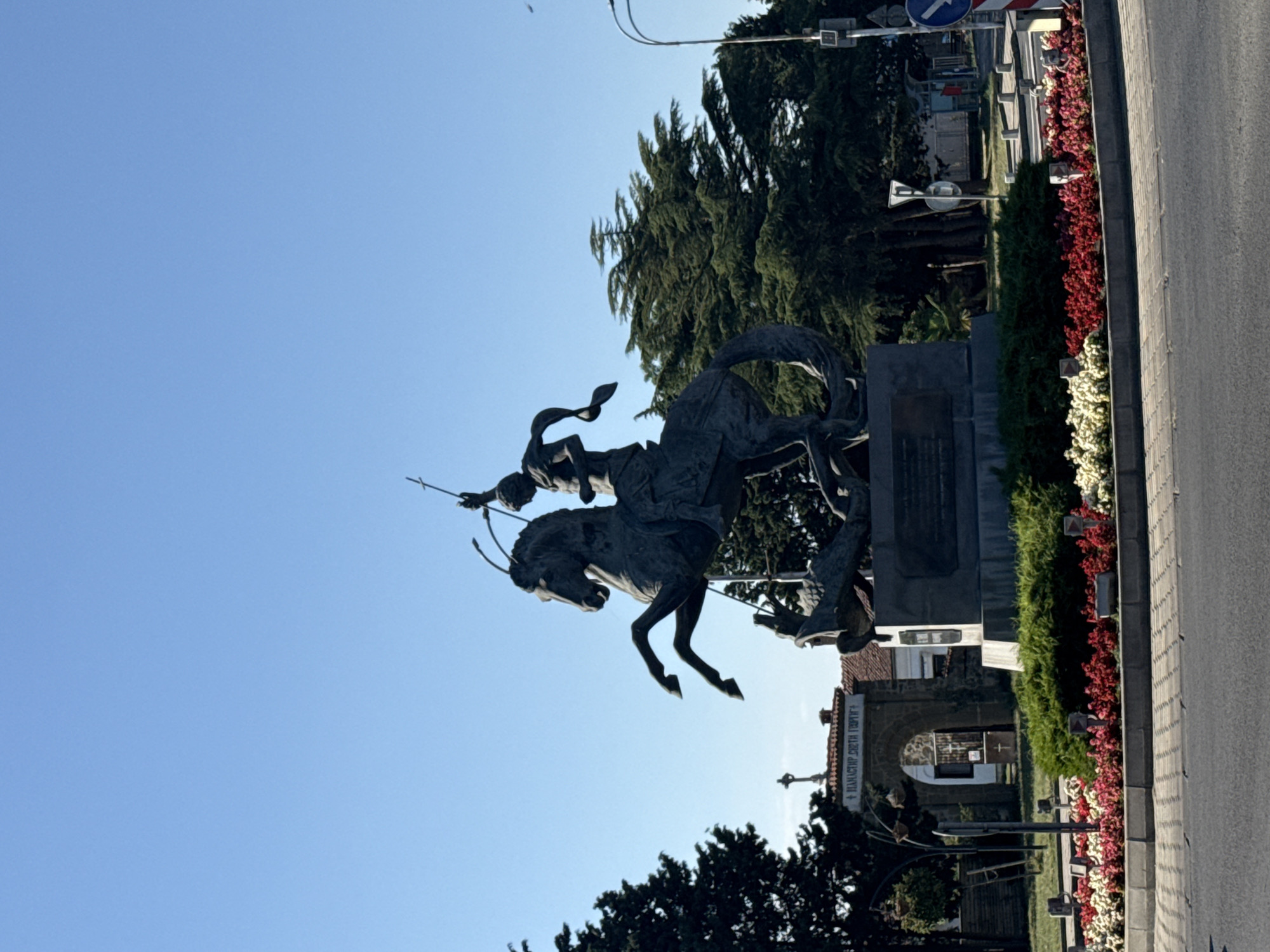 The image shows a statue of Saint George slaying a dragon, located in the city of Pomorie, Bulgaria. The statue is made of dark bronze and depicts Saint George on horseback, piercing the dragon with his spear. The horse is rearing up, and the dragon is coiled beneath it. The statue is mounted on a stone pedestal.
In the background, there is a clear blue sky, lush green trees, and a monastery with an arched entrance. The monastery appears to be an old, traditional building with red-tiled roofs. In front of the statue, there is a circular flower bed filled with red, white, and pink flowers, and neatly trimmed green bushes. The ground in front of the flower bed is paved with light-colored interlocking stones, and beyond that, a dark gray asphalt road is visible. There are also traffic signs and streetlights in the background, suggesting an urban setting.
The overall impression is one of a monument in a public space, likely a roundabout or town square, dedicated to a significant religious or historical figure. The presence of the monastery and the inscription on the archway (which appears to read "MONASTERY ST. GEORGE" in Cyrillic) strongly indicate the location as the Monastery of Saint George in Pomorie. The scene is well-lit by sunlight, suggesting it was taken during the daytime. There are no people visible in the frame.