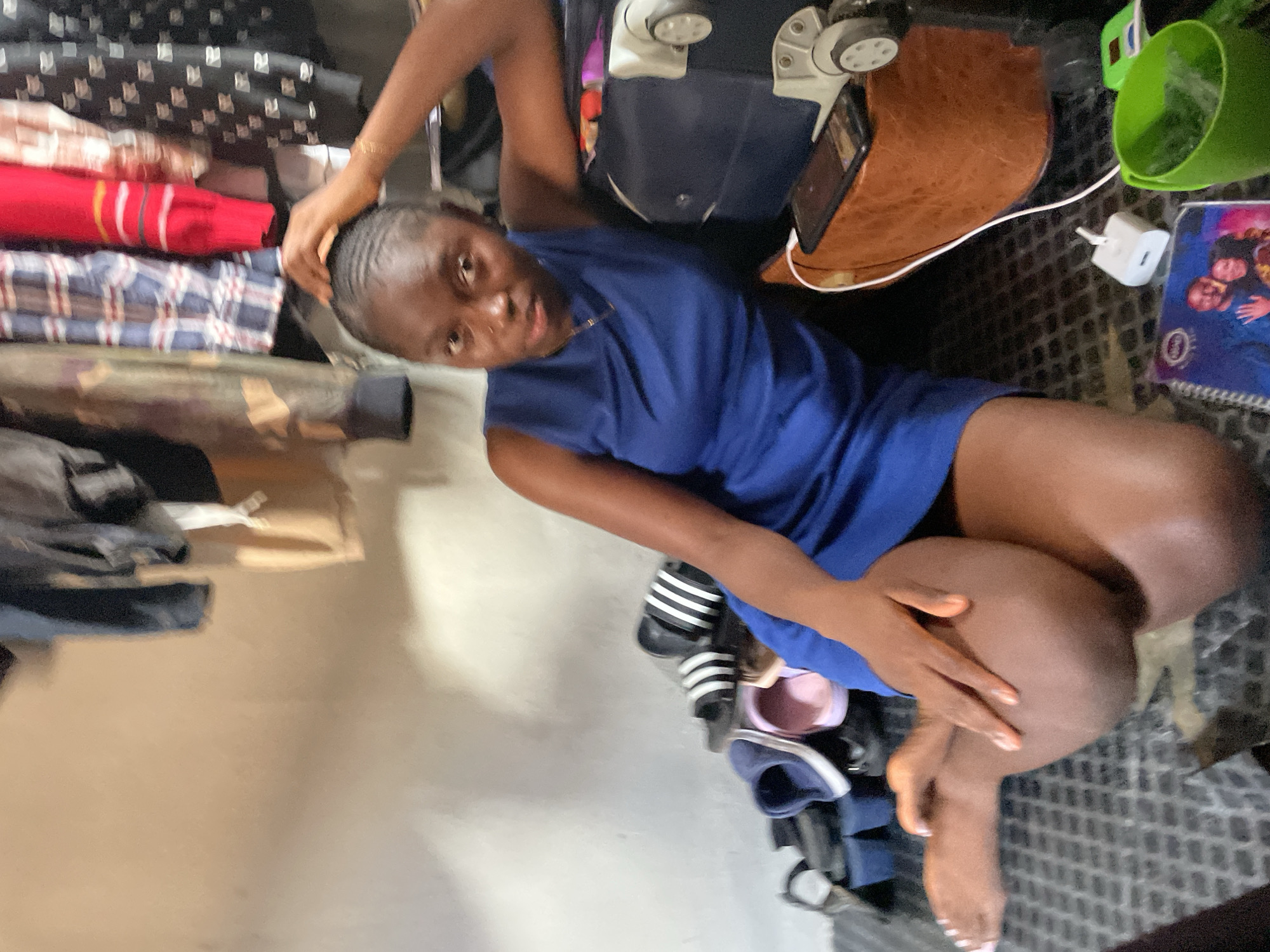 A young woman with dark skin and black hair, wearing a sleeveless blue dress, sits on the floor with her legs crossed. She is looking at the camera and has one hand on her head and the other resting on her crossed legs. Behind her, to her right, is a dark blue suitcase with wheels. Next to the suitcase is a brown bag or pouch with a smartphone placed on top of it, connected to a white charging cable. On the floor in front of the bag is a white charging brick and a green plastic cup. To the woman's left, on the floor, are several pairs of shoes and sandals, including black Adidas slides and blue and pink slip-on shoes. Behind the woman, clothes are hanging on a rack, and the wall appears to be a neutral color. The lighting suggests an indoor setting, possibly a bedroom or a storage area. The overall scene depicts a casual or perhaps transitional moment.