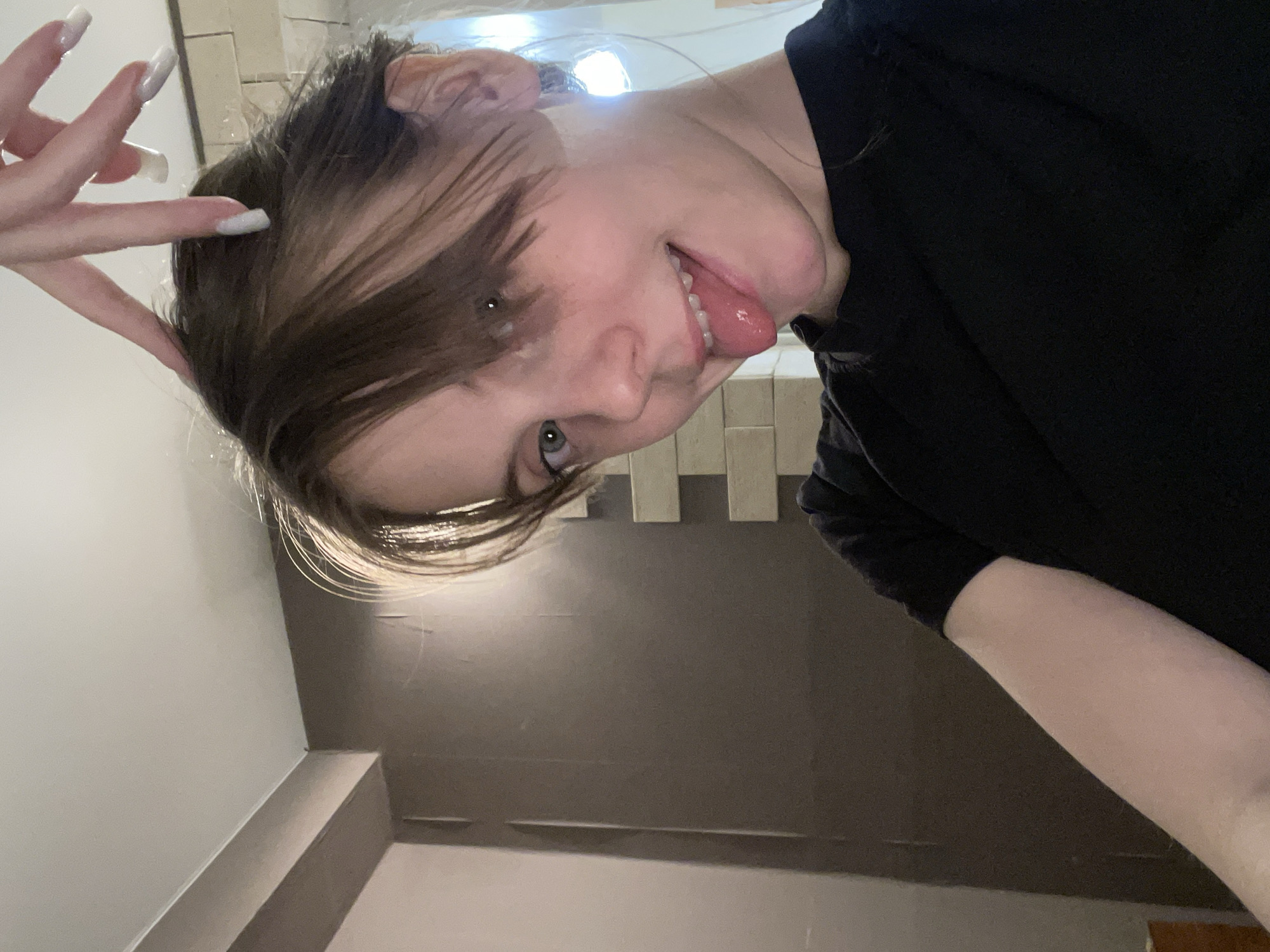 A close-up shot depicts a woman, her head tilted, sticking out her tongue. Her hair is dark with lighter strands. She is wearing a dark shirt. One of her hands is raised near her head, fingers extended. The background consists of neutral-toned walls and a tiled area near the top. The location appears to be indoors.