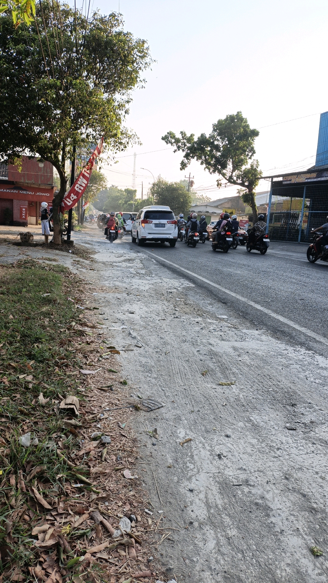This is a street-level, eye-level, slightly angled, vertical photograph taken during daylight, likely in the afternoon, in Sumbring, Indonesia. The scene depicts a busy street with several motorcycles and a white SUV. The road surface is a mix of asphalt and rougher, lighter-colored aggregate, with some debris and dried leaves on the side.
On the left side of the image, there is a grassy verge with fallen leaves and small pieces of wood. Beyond the verge, a building with a red facade is visible, featuring signs with Indonesian text, including "TEMANAN MENU JOWO" and "BUKA." A tree with green foliage stands prominently in front of this building, and a red banner with Indonesian text is hanging from its branches. A person wearing a white helmet and dark clothing is standing near the tree.
The street itself is occupied by a convoy of motorcycles, most with riders wearing helmets, and a white SUV driving in the same direction. The traffic appears to be moving at a moderate pace. In the background, more trees and buildings are visible, along with utility poles and wires. The sky is bright and clear. The overall impression is of an active urban or semi-urban environment.
