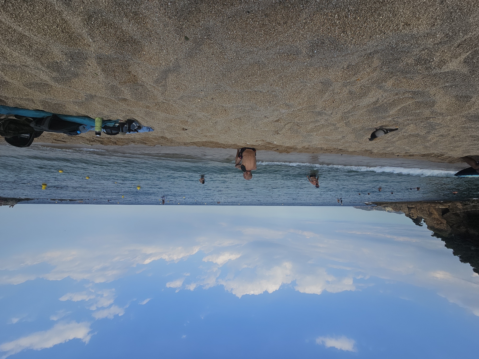 The media file depicts a sun-drenched beach scene in Blanes, Spain. The image is oriented such that the sky appears at the bottom and the beach at the top, suggesting the photo was taken with the camera inverted.
On the sandy beach, there are several bags and items of gear, including what appears to be a backpack and a blue tarp, suggesting people are preparing for or have just finished a beach activity. A single pigeon is visible on the pebbles of the beach.
The sea is a vibrant blue with small waves lapping at the shore. Several people are in the water, some wading and others swimming or engaging in water sports like paddleboarding, indicated by the presence of yellow buoys. A man in dark shorts is partially submerged in the water, facing away from the camera. Further out, other individuals can be seen enjoying the ocean. The water appears clear and inviting.
The sky is a bright, clear blue with scattered white clouds. The lighting suggests it is daytime, likely late morning or early afternoon, given the intensity of the sunlight. The presence of a rocky coastline with vegetation in the distance provides a picturesque backdrop, characteristic of the Mediterranean coast. The overall mood is relaxed and summery, typical of a vacation or day trip to the beach. No visible text is present in the media file.