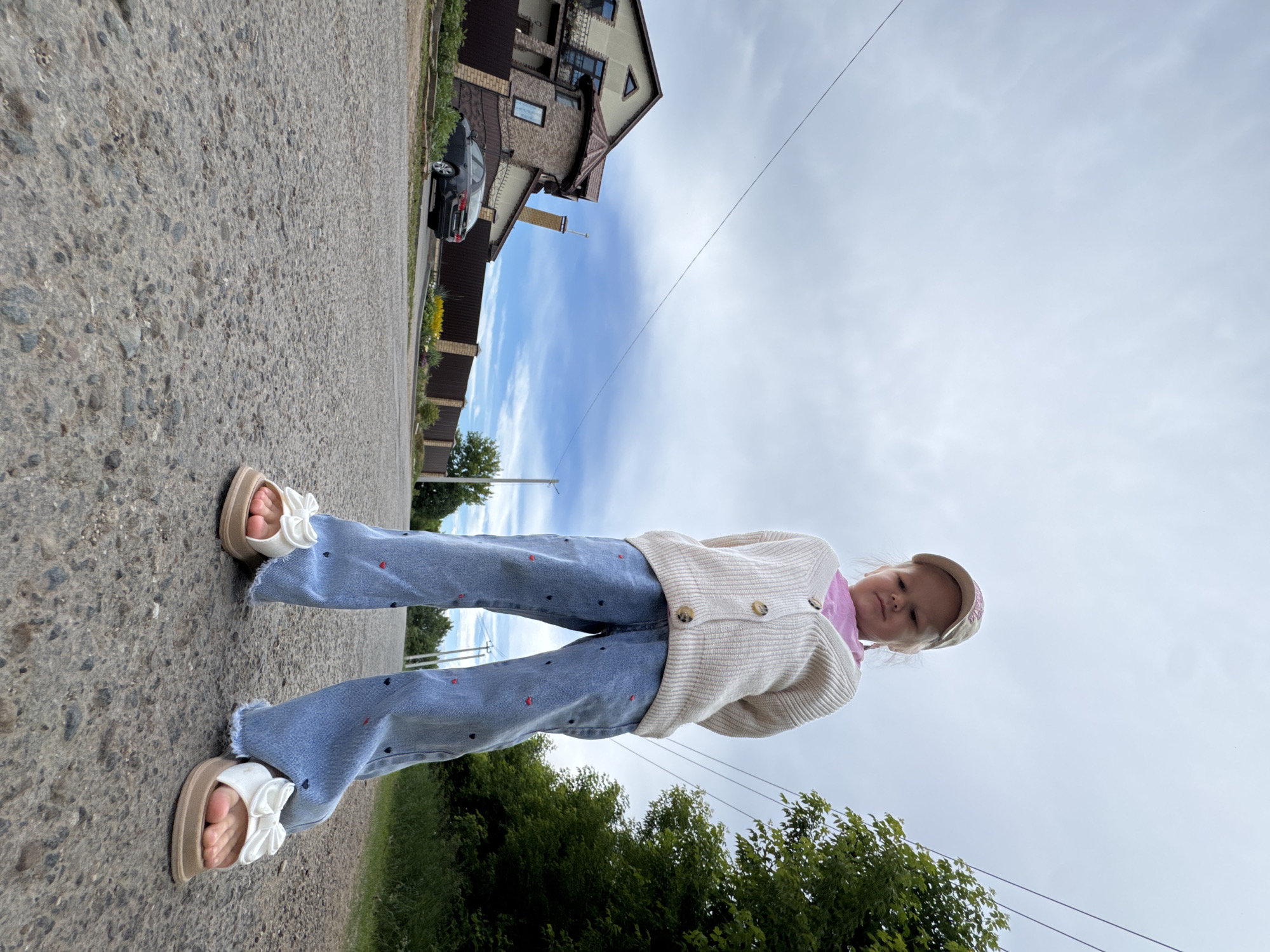 A young girl stands on an asphalt road, wearing blue jeans with red heart patterns, a beige cardigan over a pink shirt, a beige baseball cap, and white sandals with bows. She is viewed from a low angle. In the background, there is a house with a brown roof and beige siding, a dark car parked on the side of the road, a brown fence, and green trees. The sky is cloudy and gray. The location is the city of Gorniy, Russia.