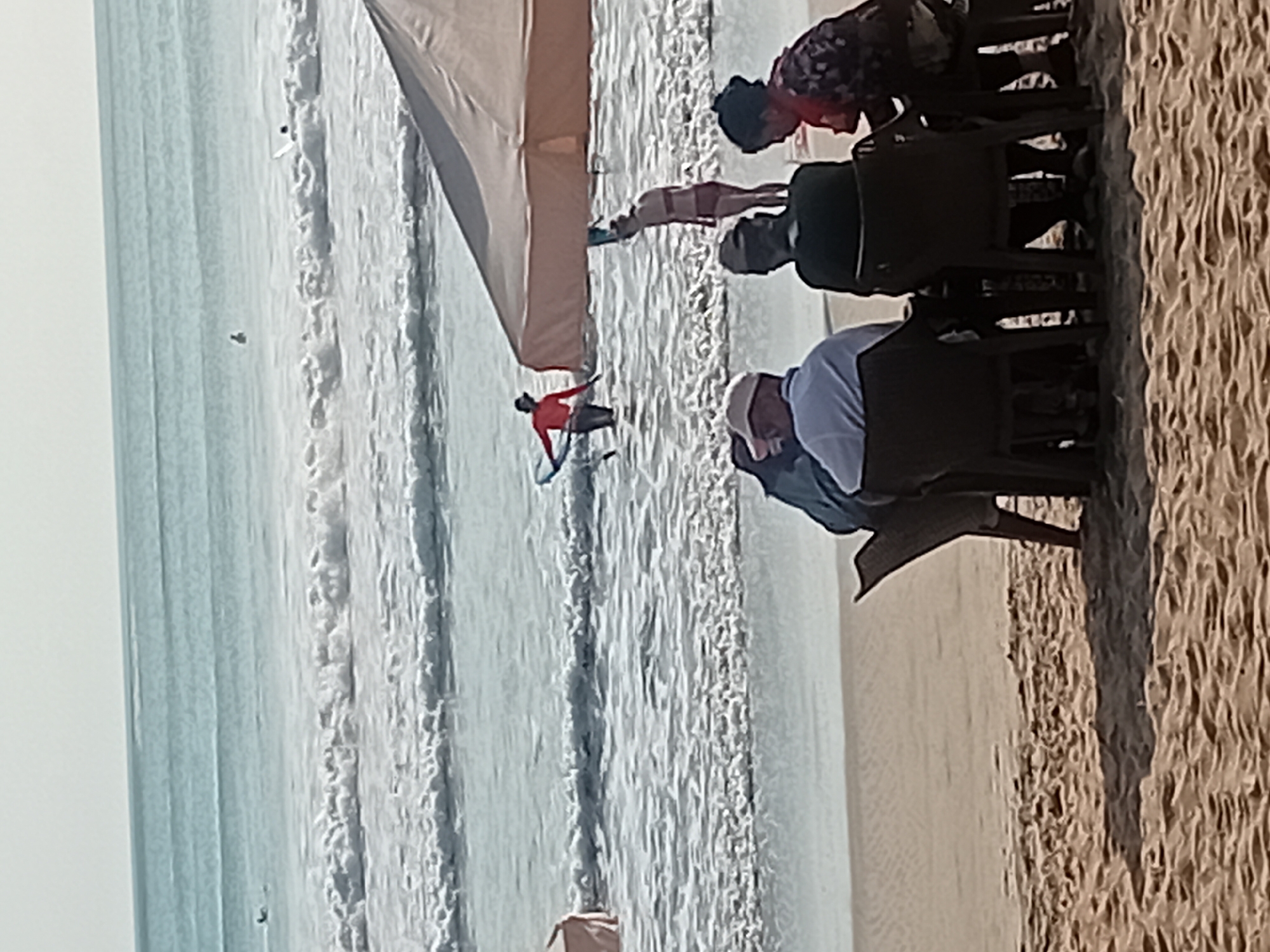 The image depicts a beach scene in Kuta, Indonesia. In the foreground, four individuals are seated at a table on the sand, partially shaded by a tan umbrella. Two of them are facing away from the camera, one wearing a white hat and a blue shirt, and the other wearing a dark cap and a dark shirt. The third person, on the right, faces the table and wears a patterned top and a red scarf. The fourth person is partially visible from behind on the far left. In the background, the ocean is visible with gentle waves breaking on the shore. Several individuals are in the water, with one person in an orange shirt holding a surfboard, appearing to be in shallow water. Another person, in a bikini, is walking with a surfboard under their arm. The sky is overcast.