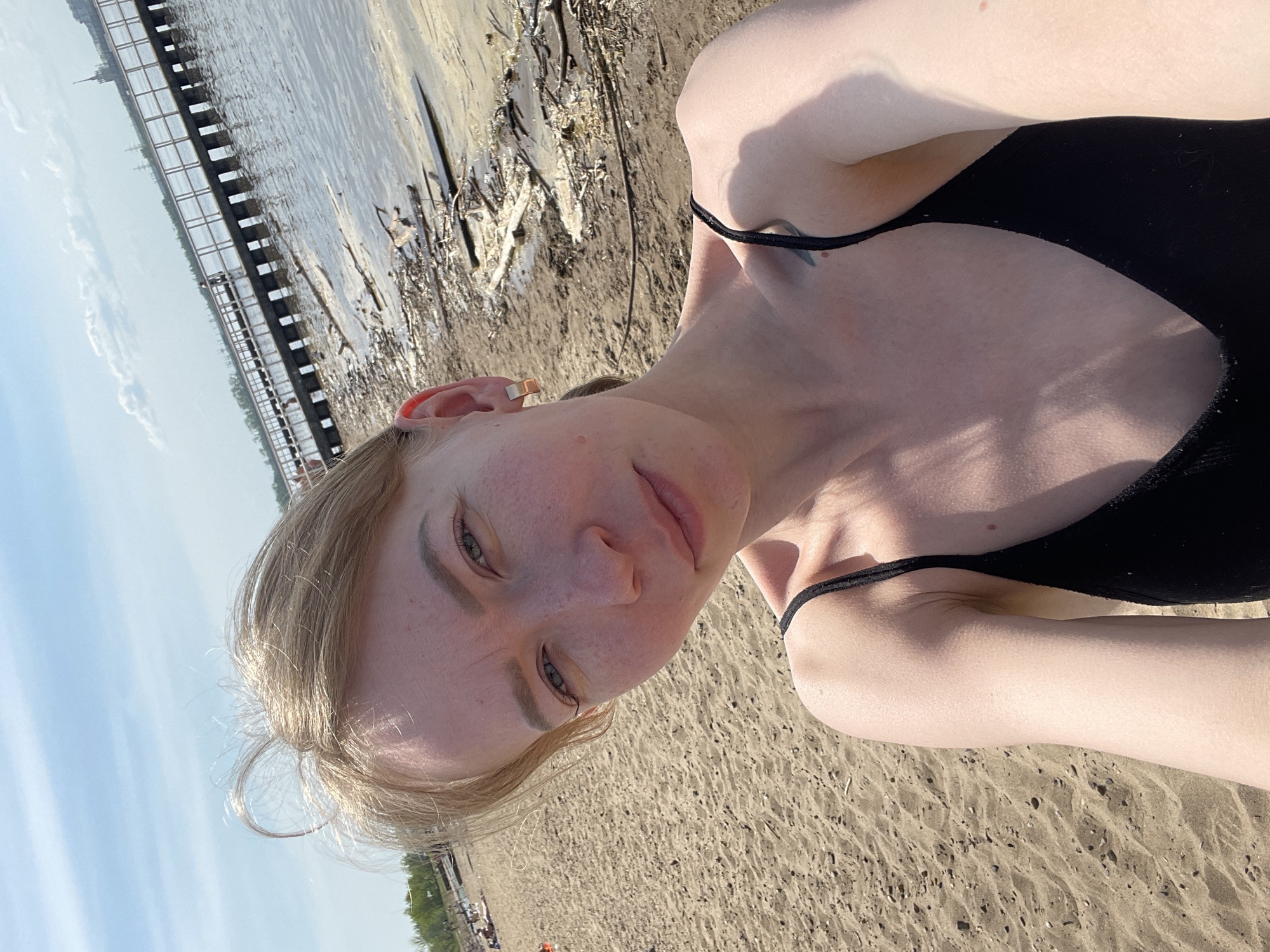 A young adult woman with light blonde hair and fair skin is taking a selfie at a beach along the Kama River in Perm, Russia. She is wearing a black top. The beach consists of sand. In the background, there's a view of the river and a bridge extending over it. The sky is light blue.