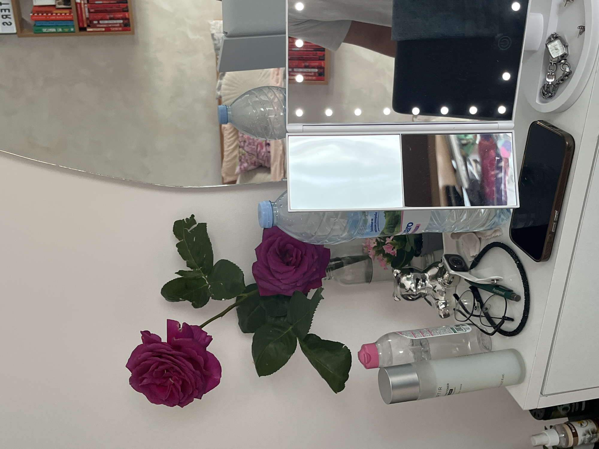 The image shows a vanity table with a variety of items. On the left side, there is a mirror reflecting the interior of a room. The reflection includes a shelf with books, pillows, and the back of a lighted makeup mirror.
On the right side of the table are a lighted makeup mirror, a water bottle, and a cosmetic organizer containing a watch and earrings. There is also a cell phone on top of the organizer. Near the front of the table are a small decorative silver figurine, a pair of glasses, a pen, a container of liquid with a pink lid, and a bottle of toner.
Three roses, two dark pink and one lavender, are arranged at the front of the table.
The image doesn't contain any information indicating the location in Blagoevgrad, Bulgaria.