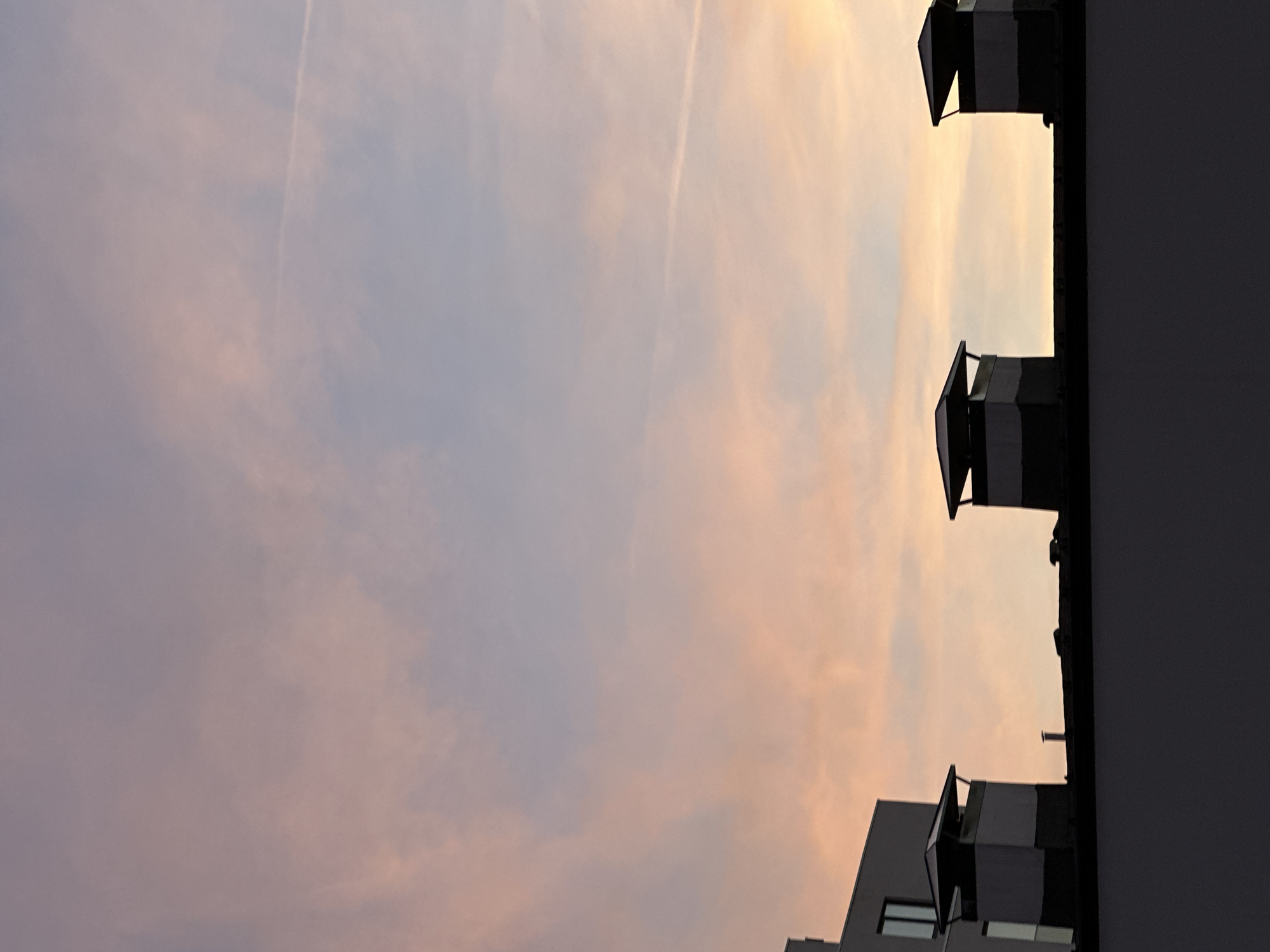 The image shows a view of the sky at sunset from Sofia, Bulgaria. The sky is filled with clouds that are colored in shades of pink, orange, and blue. There are several buildings in the foreground, including apartment buildings. The buildings are silhouetted against the sky, and their windows are visible. The photo is taken from the ground, looking up at the sky and buildings. The angle makes the sky look large and expansive. There is also the contrail of an airplane in the sky.