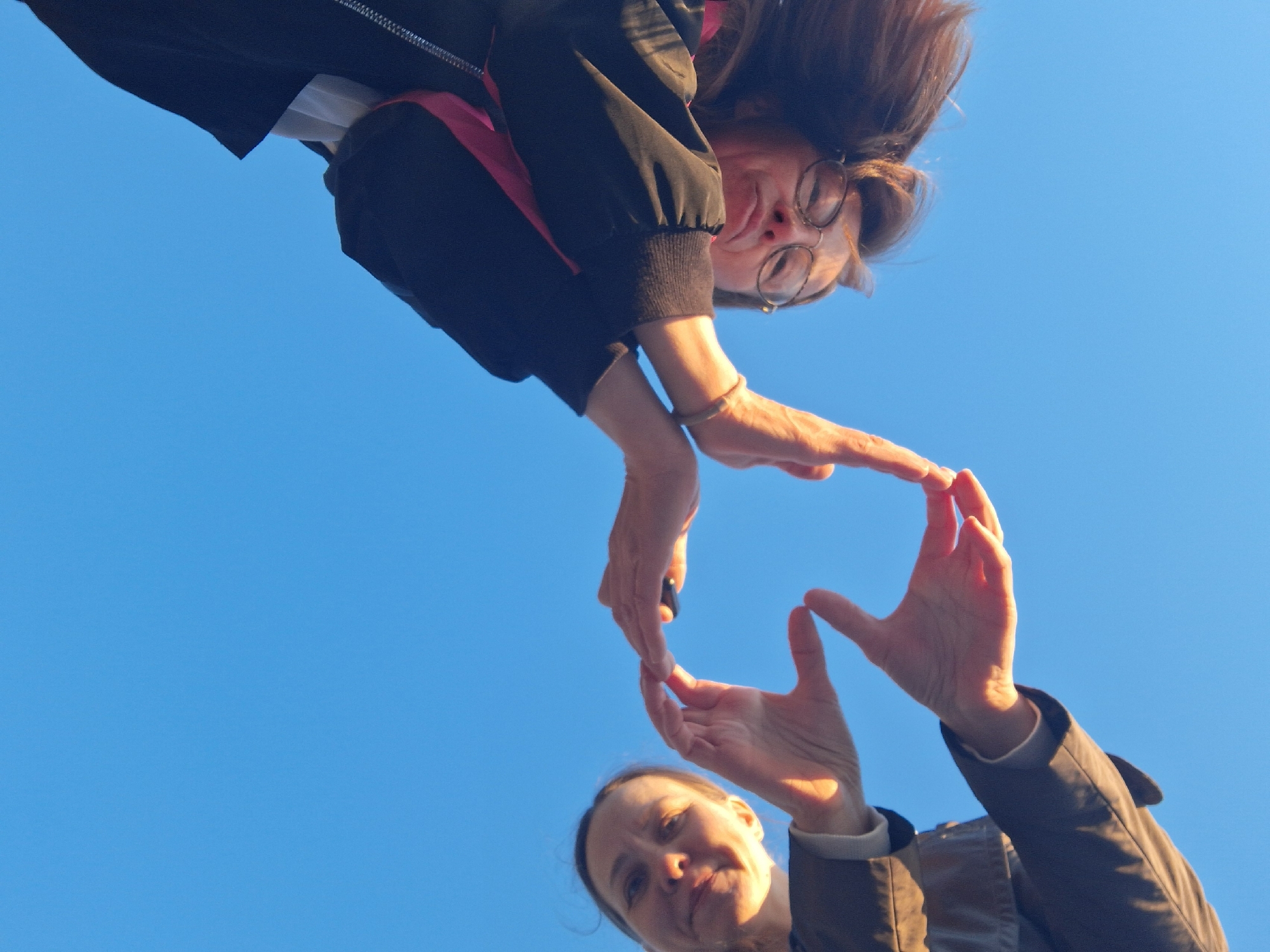 From a low-angle perspective, two individuals are pictured against a clear blue sky. The person above is wearing glasses and a jacket with a pink accent. The person below is wearing a brown jacket. The two people's hands come together to form a heart shape. The location is Chaikovsky, Russia.