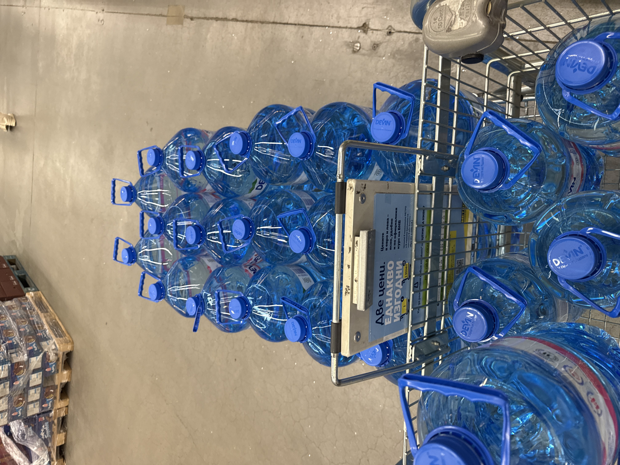 The image shows a shopping cart full of large, blue water bottles with "Devin" labeled on the blue caps. The water bottles are stacked tightly in the cart, and the cart is parked on a grey concrete floor. An advertisement is affixed to the side of the cart with text in Bulgarian: "Две цени. Еднакво Изгодни," and below it lists a price of 1.95583. Pallets loaded with goods are visible in the background. The scene is most likely within a large supermarket or warehouse-style store in Burgas, Bulgaria.