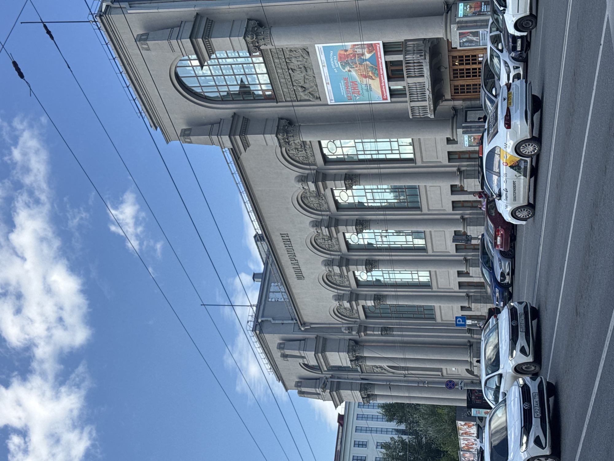 The image shows a wide street in front of a large, ornate grey building with a sign that reads "ФИЛАРМОНИЯ" (Philharmonic). This is the Sverdlovsk Philharmonic Hall located in Yekaterinburg, Russia. Several cars are driving on the street and parked in front of the building. The sky is blue with scattered white clouds. There are trolley wires visible in the sky. A large poster is displayed on the building, advertising an event. The architecture of the building is classical with columns and arched windows. The overall scene depicts a sunny day in a city.