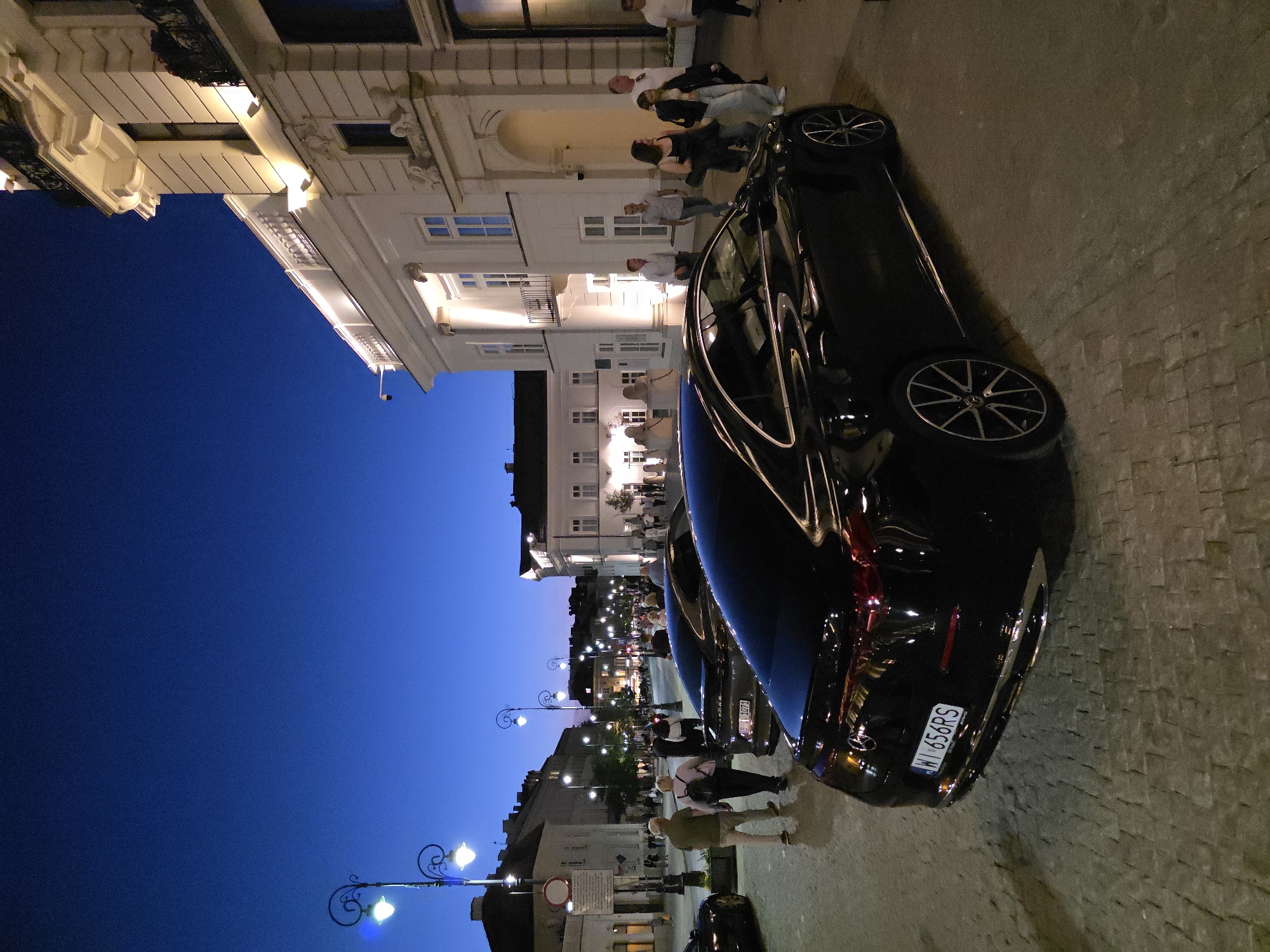 Captured at night, the image shows a street scene in Warsaw, Poland. The sky is a deep blue. The area is illuminated by ornate street lamps and building lights. Along the street, people are strolling, some are standing in groups and talking. Parked along the side of the street is a black Mercedes-Benz with the license plate "W 656RS". The architecture of the buildings is characterized by classical facades. The ground is paved. The setting appears to be a historic district or main thoroughfare.