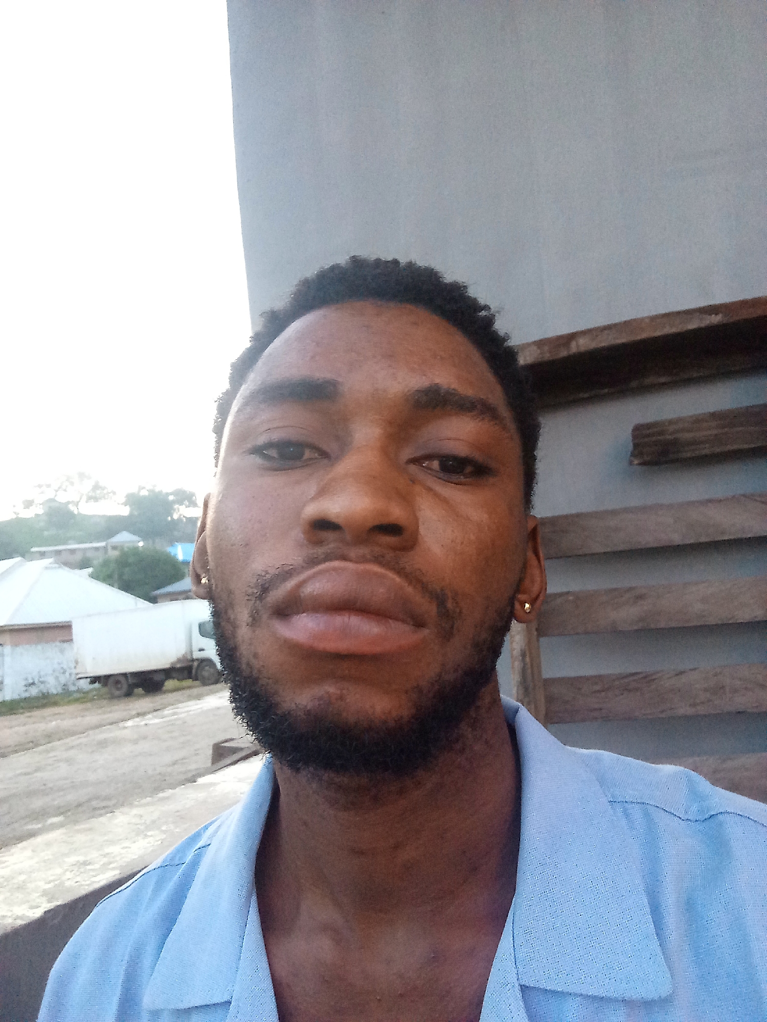 A head and shoulders, selfie-style, portrait of a Black male is presented. The man's face is turned towards the camera, his mouth is slightly open, and his eyes are looking forward. He has short, dark, curly hair, a goatee, and is wearing a light blue collared shirt with the top buttons undone. A small gold earring is visible in his right ear.
The background is blurred and consists of what appears to be an outdoor setting. To the left, there is a white building with a white truck parked in front of it. Beyond the building, trees and other structures are faintly visible, suggesting a town or village environment. To the right of the man, there is a grey wall with wooden planks attached to it in a horizontal arrangement.
The overall lighting suggests it is daytime, possibly late afternoon or early morning, given the bright glare from the left side of the image. The quality of the image is moderate, with some grain and a slight blur.
This image is likely not in Lokoja, Nigeria, as the scene does not contain any identifiable landmarks or context that would place it there. The environment depicted is generic and could be found in many locations.