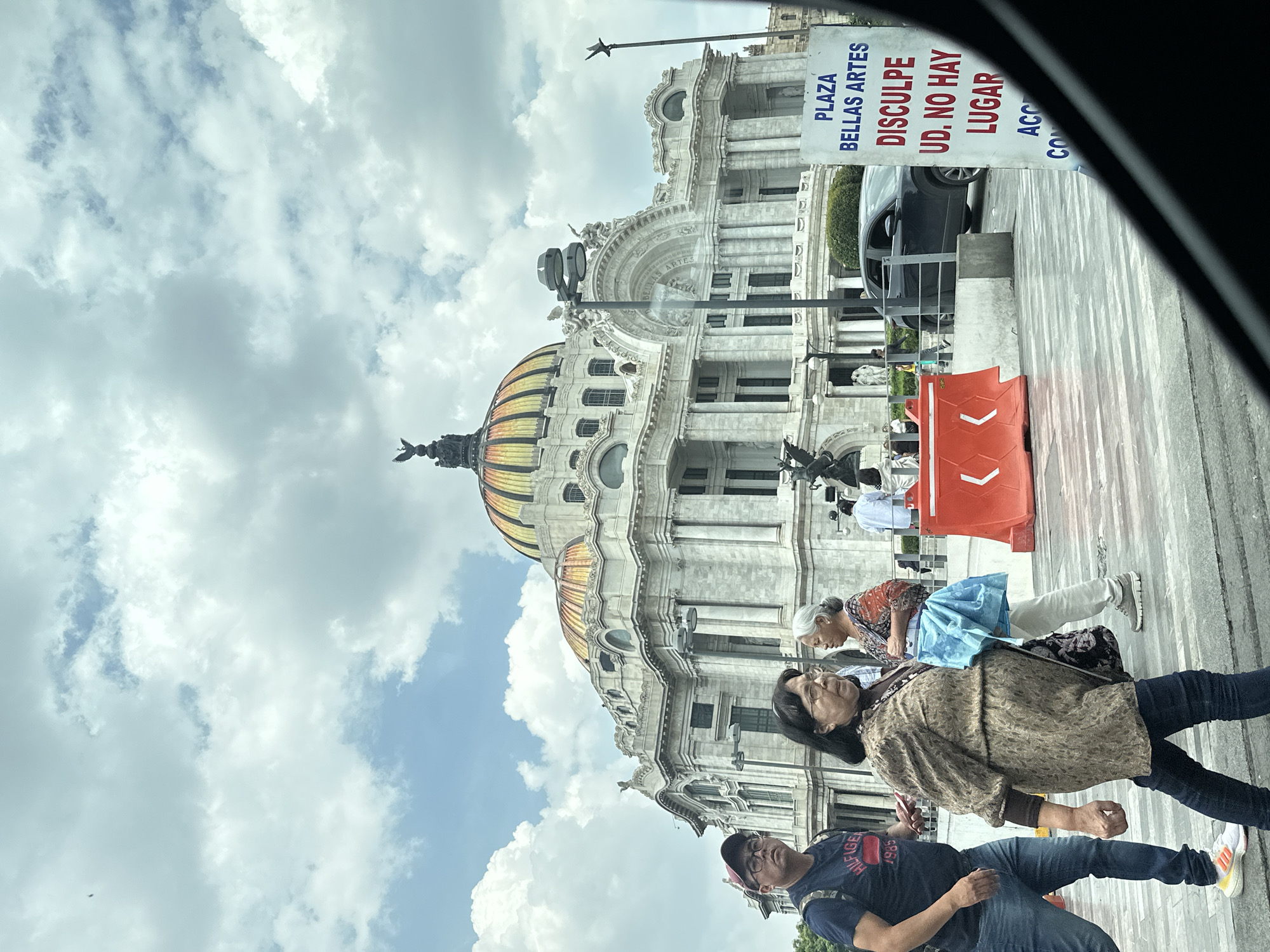 A daytime, eye-level shot captures the exterior of the Palacio de Bellas Artes in Mexico City. The grand building, made of white marble, features a prominent dome with yellow and gold accents and is adorned with sculptures. In the foreground, a group of people are walking on a paved area, with some individuals in focus and others blurred due to motion. A man in a blue t-shirt and jeans walks on the left, followed by a woman in a patterned brown jacket and dark jeans. Another woman in a patterned top and white pants walks behind her, carrying a light blue bag. To the right of the building, a white sign with red text reads "PLAZA BELLAS ARTES DISCULPE UD. NO HAY LUGAR ACC CO". A black car is parked nearby, and orange traffic barriers are visible in the midground. The sky is filled with white and gray clouds.