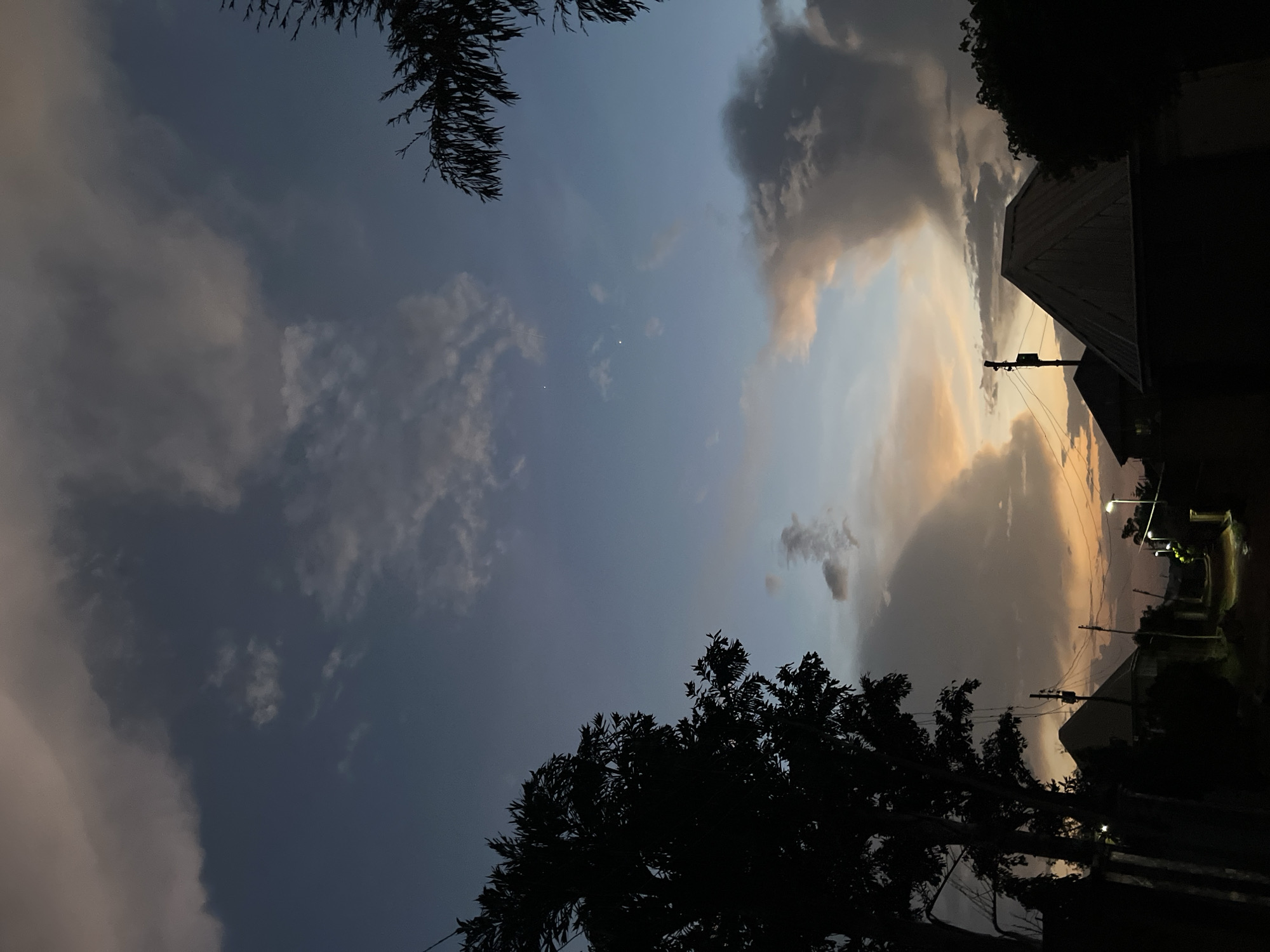 This vertically oriented image captures an outdoor scene at twilight, likely dusk, in a residential or semi-urban setting like Jos, Nigeria. The dominant subject is the sky, which fills most of the frame with a dramatic display of clouds and colors. The sky transitions from a deep, dusky blue-grey in the upper and central parts to warmer golden and peach tones along the right side, where the last light of the day lingers. Various cloud formations are visible, ranging from dark, heavy masses to lighter, more ethereal wisps that catch the sky's soft glow. Two distinct, bright white dots, possibly stars or planets, are discernible in the central-left area of the sky.
Silhouetted against the vibrant sky are several elements of the landscape. In the top left, the dark, fringed fronds of a palm or similar tree extend into the frame. Along the bottom, a dense mass of tree canopies forms a jagged silhouette. On the right side, the angular shapes of building roofs, likely residential structures, are sharply outlined against the lighter horizon. A utility pole with multiple overhead wires runs vertically on the right, connecting to the unseen infrastructure. Further along the right edge, what appears to be a street or pathway is dimly lit by several glowing streetlights, casting a warm, yellow light onto the ground and suggesting the deepening evening. No people are visible, and no text is present in the image. The scene evokes a quiet, peaceful atmosphere as day transitions into night.