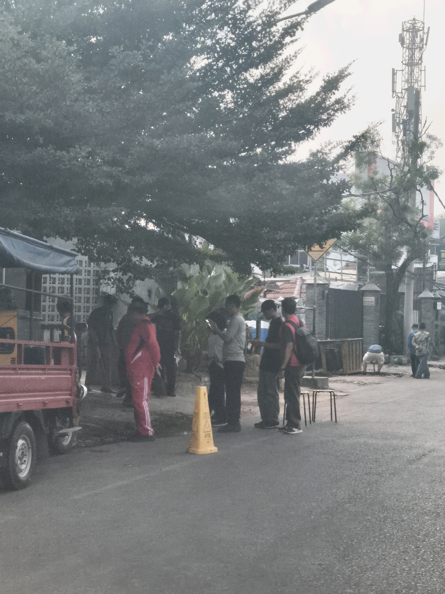 In Palembang, Indonesia, a group of individuals are gathered on a street. A red truck with a flatbed is parked on the left side of the image. In the foreground, a yellow traffic cone is placed on the asphalt. Several men, some wearing casual clothing and others in more formal attire, are standing and conversing. One individual in the middle is looking at their phone. Further in the background, a cell tower stands tall against a hazy sky, with lush green trees partially obscuring the view. Buildings and a fence are also visible in the background, suggesting an urban or semi-urban environment. The overall lighting indicates it is daytime, possibly late afternoon or early morning due to the soft light.