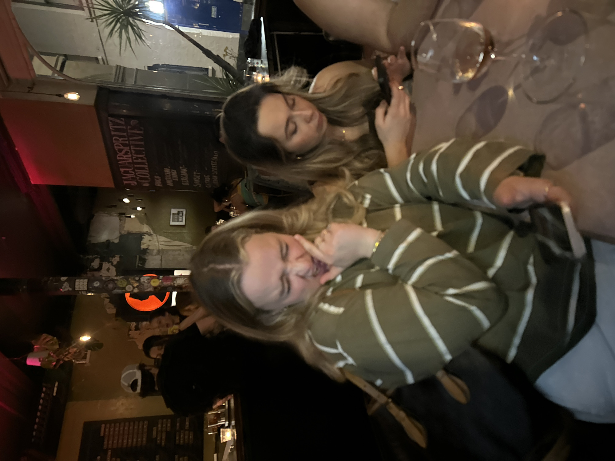 The image shows two women at a table with wine glasses. The woman closer to the camera is blonde and wearing a green and white striped shirt. She has her hand up to her mouth. The other woman is looking at her phone. They are at a table at the Jaguar Spritz Collective in London, United Kingdom.