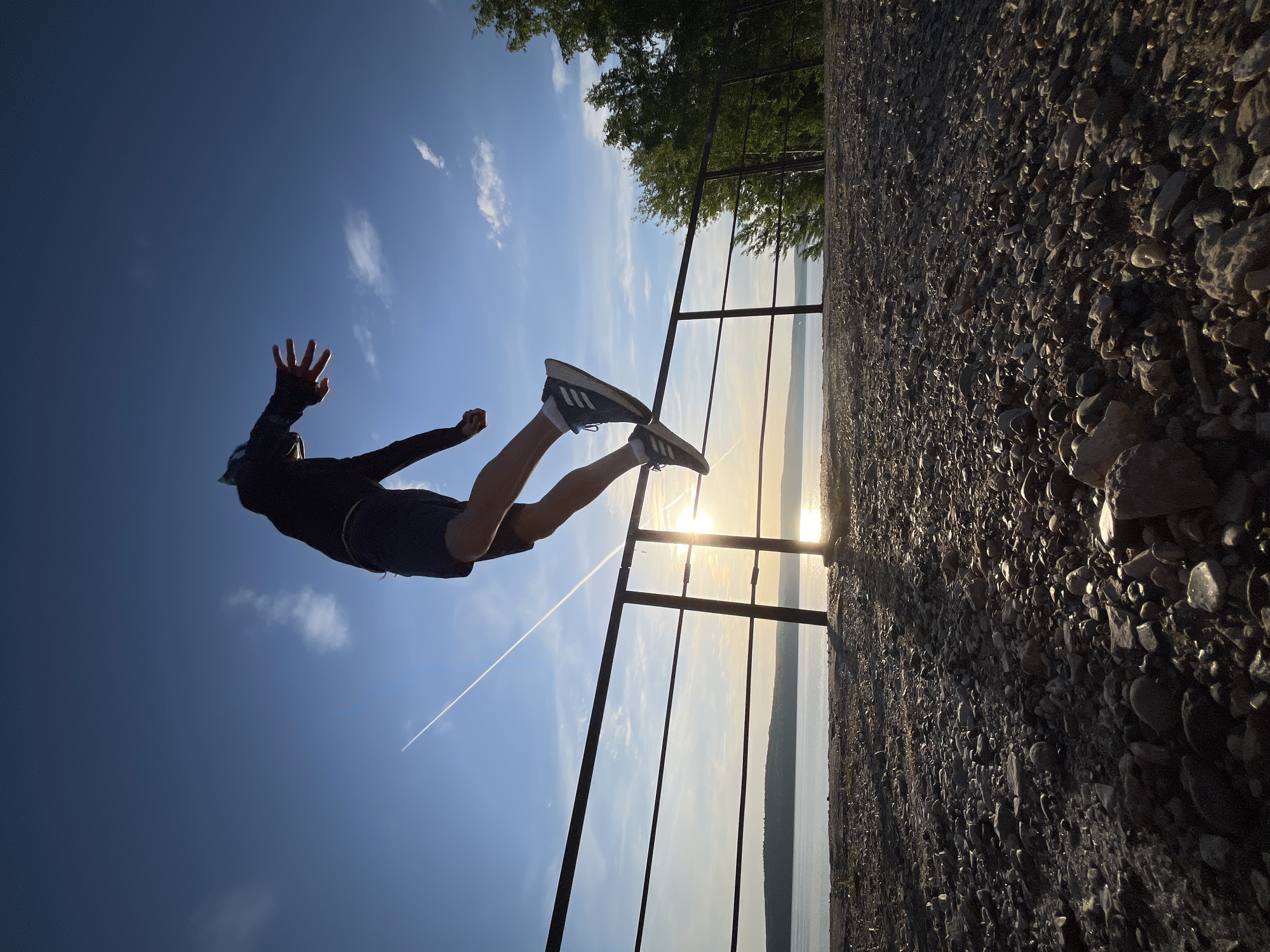 A person is suspended horizontally in the air, legs extended towards the right, arms outstretched to the left, in front of a metal railing. The ground consists of small rocks and pebbles, located to the right. Beyond the railing, a body of water is visible with sunlight reflecting off its surface. The sky is blue with wispy clouds, with a contrail from an aircraft visible. The image appears to be taken from a low angle, exaggerating the height of the person. The location is likely a viewpoint or elevated platform in Chaikovsky, Russia, overlooking a body of water.