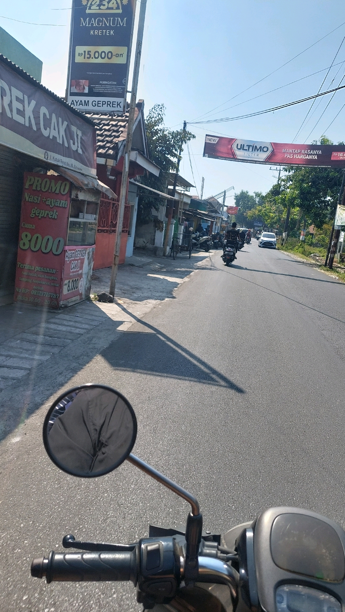 The image captures a street scene in Sidoarjo, Indonesia, from the perspective of someone riding a motorcycle. In the foreground, the handlebars and a rearview mirror of the motorcycle are visible. The road ahead is paved and appears to be a two-lane street with light traffic. On the left side of the road, there are several small shops and stalls. One stall prominently displays a red sign with "PROMO" in large white letters and text advertising "Nasi + ayam geprek" for "8000." Above this stall, a larger sign reads "REK CAK JI." Further down the street, another sign advertising "MAGNUM KRETEK" and "AYAM GEPREK" is visible, along with a banner advertising "ULTIMO FILTER." Several motorcycles and a white car are present on the road. The environment is sunny with a clear blue sky. The overall scene depicts a typical commercial street in an Indonesian town, characterized by small businesses, motorcycles, and a lively atmosphere.
