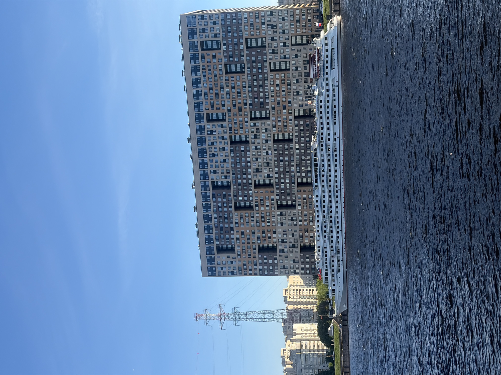 A large, modern apartment building with a complex facade of varying window arrangements and colors stands on the bank of a dark blue body of water. A long, white passenger ship is docked in front of the building. To the left of the apartment building, a tall, lattice-style power transmission tower is visible against a clear blue sky. Further left, a row of more traditional, multi-story apartment buildings can be seen. The scene appears to be an urban waterfront. The location is the city of Shushary, Russia, specifically along the Neva River.