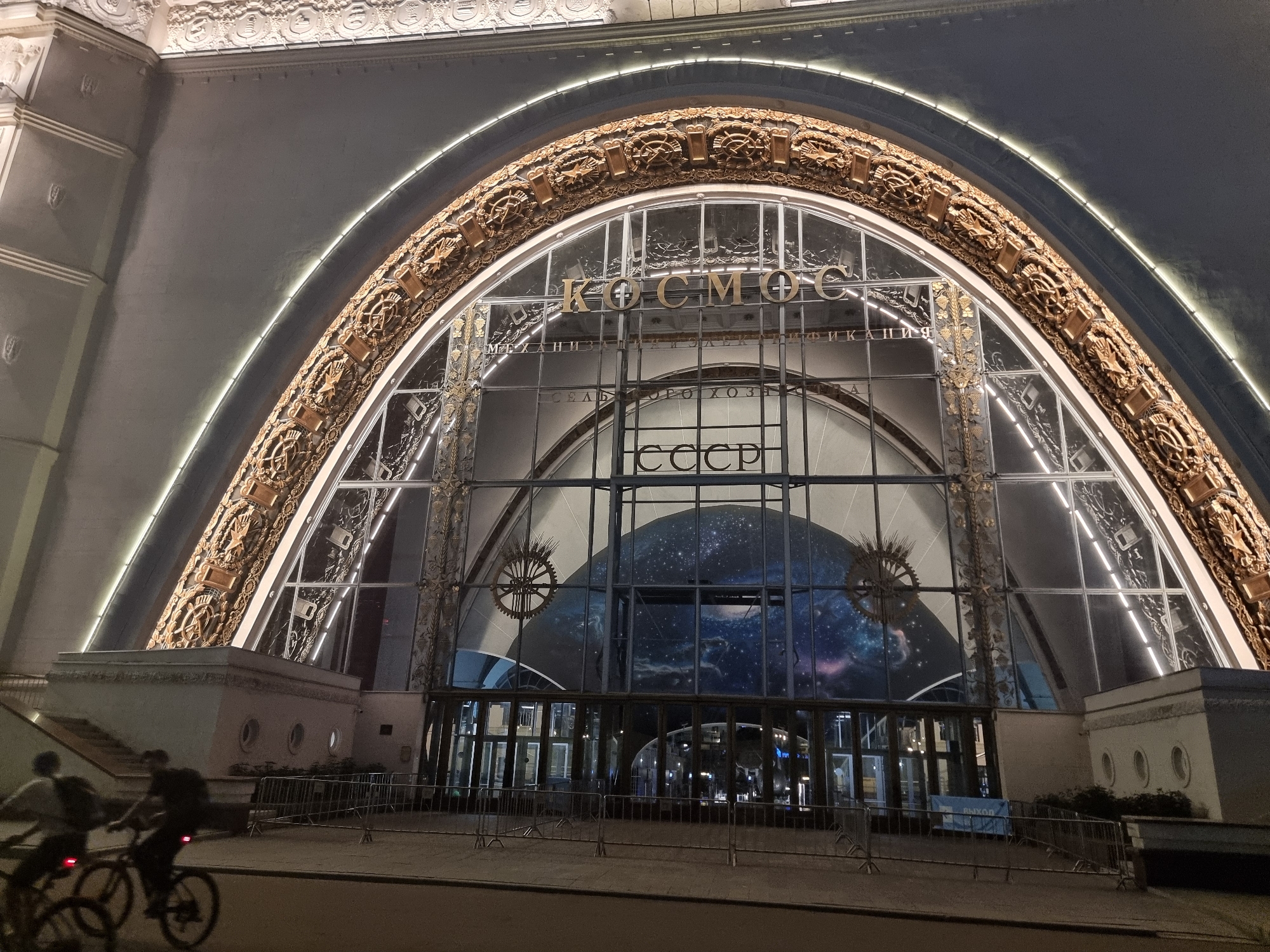 The image shows the exterior of the Moscow State Space Museum (also known as VDNKh's Cosmonautics Museum or the Monument to the Conquerors of Space). The building features a large, arched glass facade with ornate golden detailing. Above the entrance, the word "КОСМОС" (Cosmos) is visible in large letters, and below it, "СССР" (USSR) is also displayed. Inside the glass facade, a large spherical display resembling a planet or galaxy is visible. The exterior is illuminated by lights along the arch and from within the building. In the foreground, two people are riding bicycles on a paved area, with metal barriers set up along the perimeter. The scene appears to be at night, with the lighting creating a dramatic effect on the architecture.