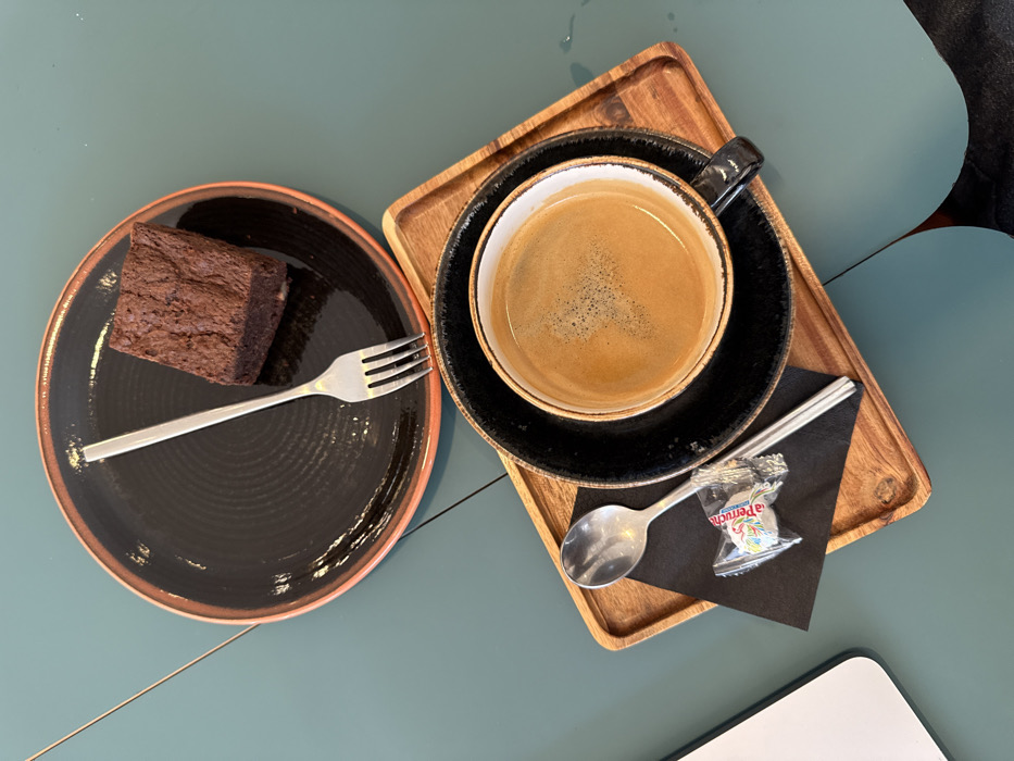 An overhead view captures a dessert and coffee arrangement on a teal-colored table in Le Havre, France. In the upper center, a round black plate with an orange-brown rim features a textured surface and holds a single rectangular chocolate brownie. A silver fork rests diagonally across the plate, tines pointing towards the upper left. Below this, a black ceramic coffee cup with a lighter interior, containing a dark brown liquid topped with light brown crema, sits on a matching black saucer. This coffee setup is positioned on a small, rectangular wooden tray with raised edges. Adjacent to the coffee cup on the tray, a silver spoon lies on a folded black napkin, next to a clear plastic-wrapped candy, identifiable as a "La Perruche" sugar cube. A portion of a white object, likely a laptop, is visible at the far left edge of the teal table. A subtle diagonal seam is present on the table's upper left surface, and a faint water mark or reflection is visible on the lower right of the table.