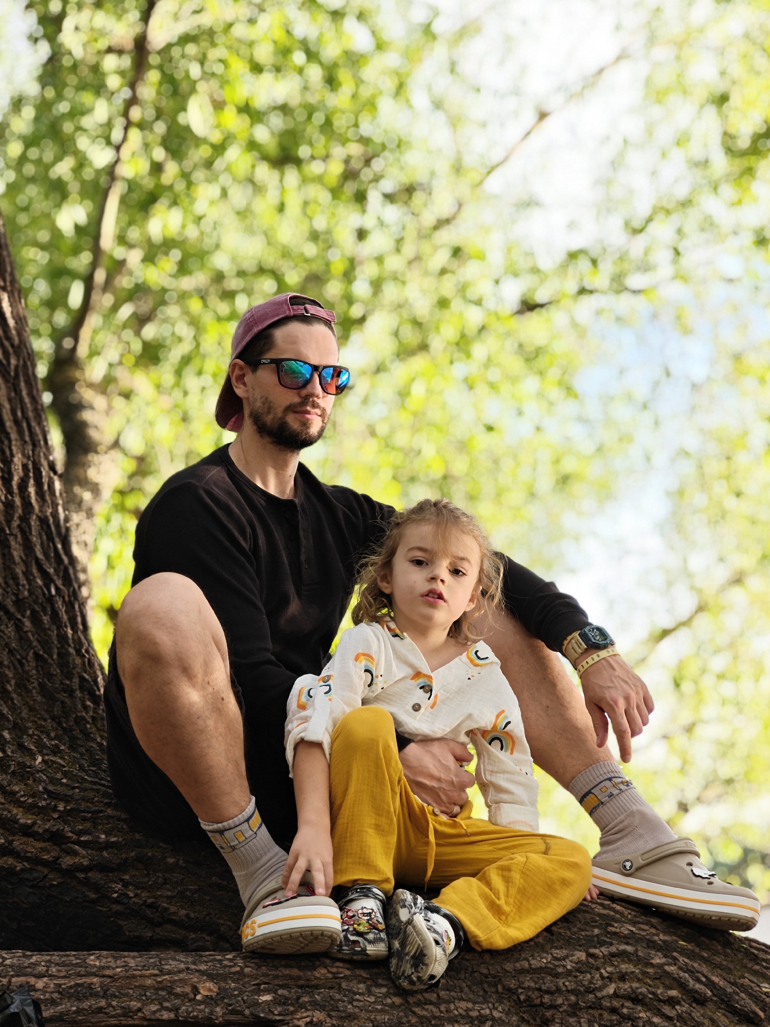 A man and a young child are sitting on a large tree branch. The man has a beard, wears a burgundy baseball cap, dark sunglasses, a black long-sleeved shirt, and dark shorts. He has a watch on his left wrist and wears socks with Crocs shoes. The child, with light brown hair, wears a white button-up shirt with colorful rainbow designs and yellow pants, with Crocs shoes on their feet. The tree branch is thick and textured. The background is out of focus and full of green and yellow foliage, suggesting a park or wooded area. The image was taken in the city of Khimki, Russia.