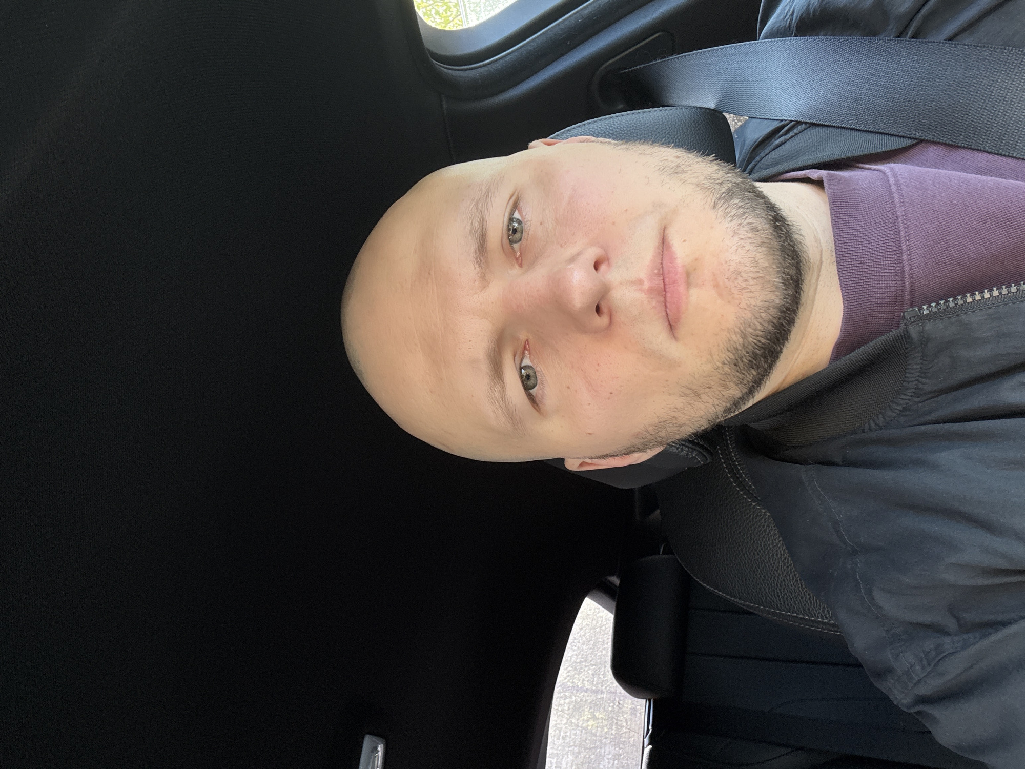 A bald man with green eyes and a beard is sitting in a car wearing a seatbelt. He is wearing a purple shirt under a black jacket. The interior of the car is black. The location cannot be determined from the provided image.
