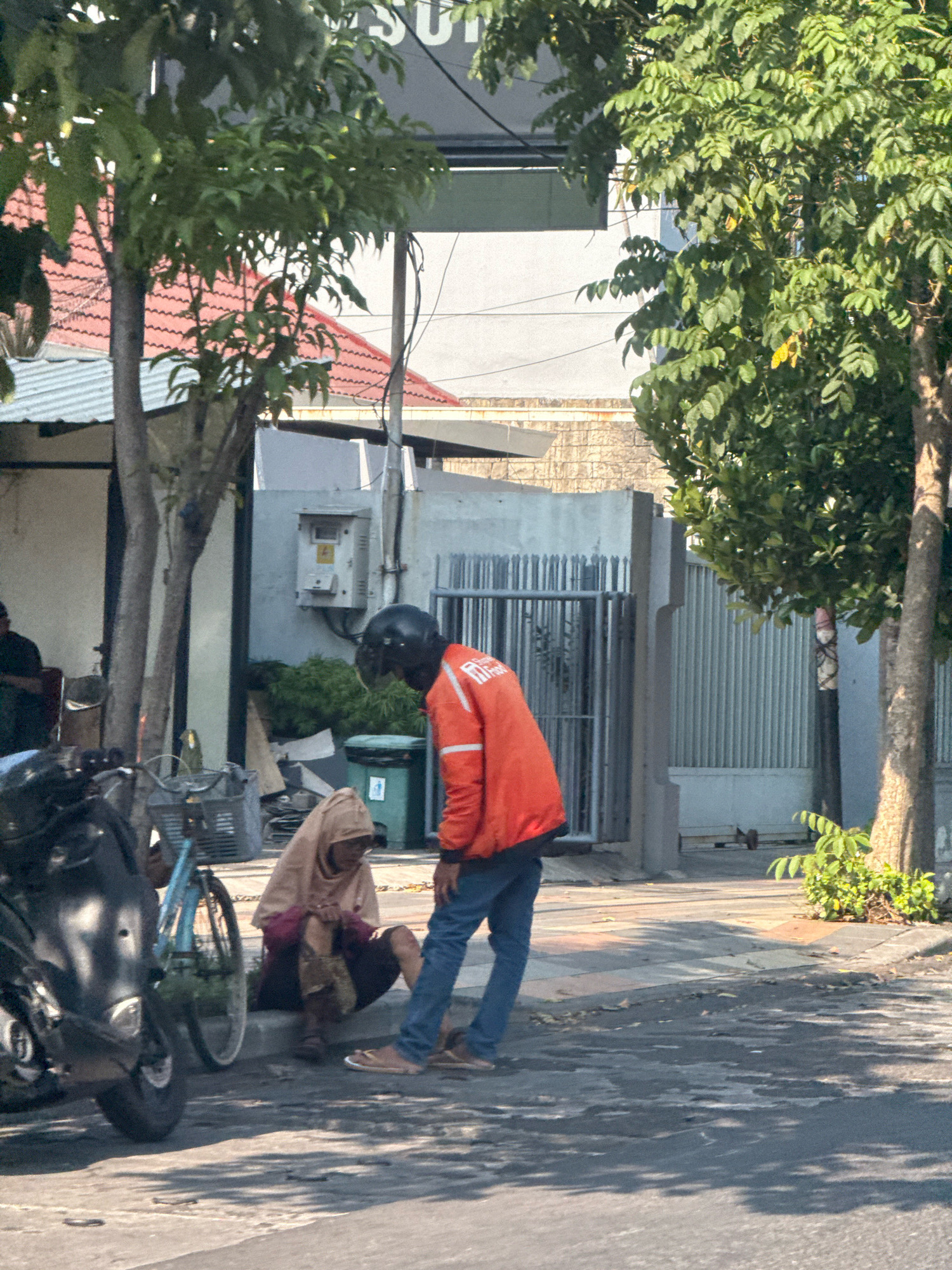 In Surabaya, Indonesia, on a sunny day, a person wearing a helmet and an orange jacket stands on a sidewalk next to a woman who is sitting on the curb. The woman is wearing a tan hijab and a dark skirt. She appears to be holding some items in her lap. In the foreground, a motorcycle is parked on the left side of the image, and a bicycle is behind it. In the background, there are buildings, trees, and a metal fence. The scene appears to be a street or a roadside area.
